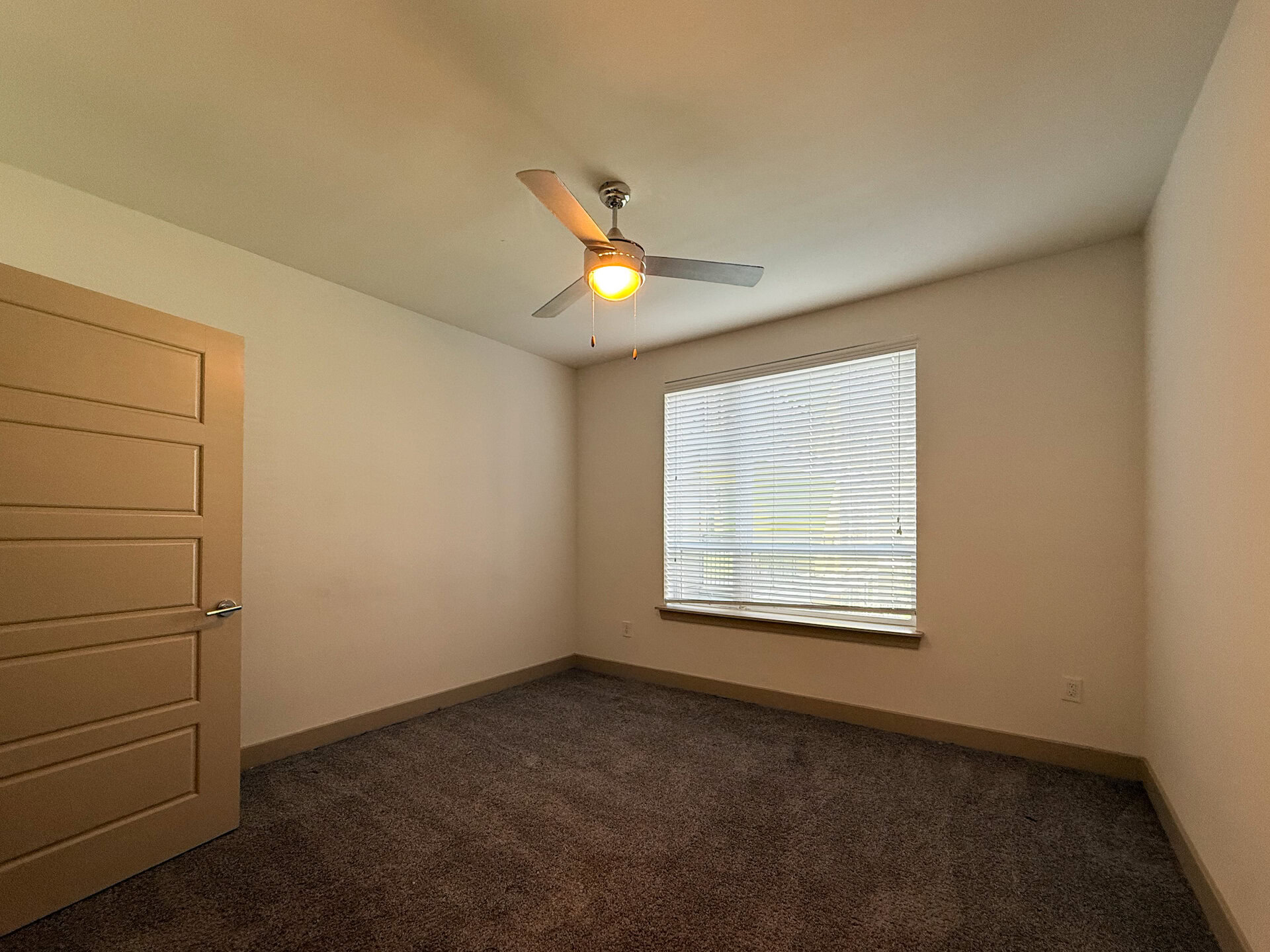 A spacious bedroom with carpet and horizontal blinds allows natural light to enter, while a beige door with a modern design is visible on the left side of the image. The overall atmosphere is bright and minimalistic.