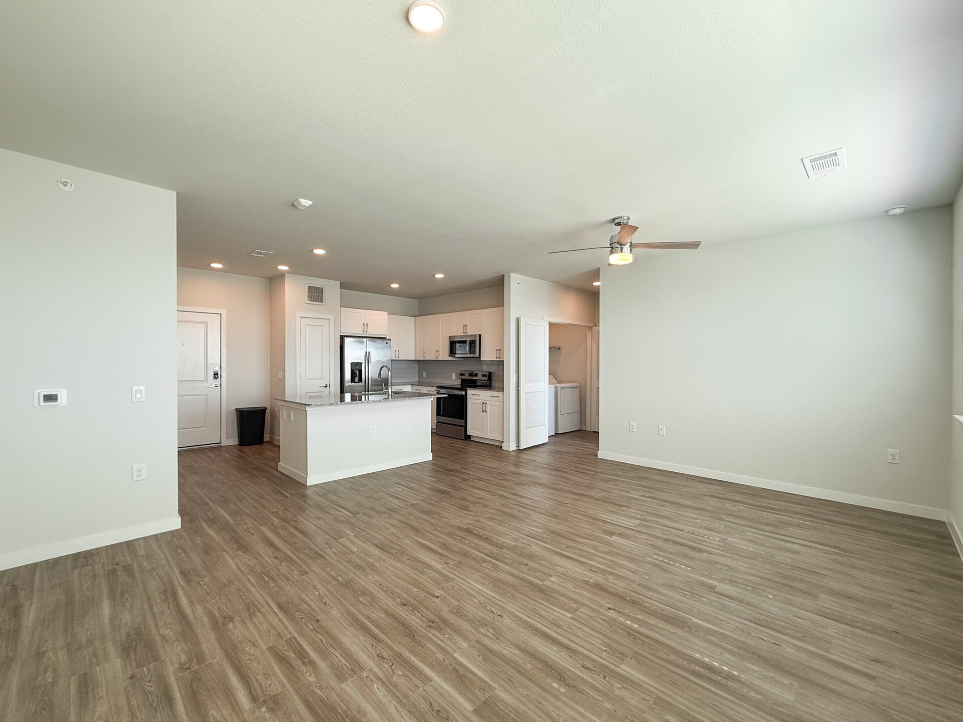 A spacious, modern living area features light-colored walls and a wood-like laminate floor. On the left, a front door leads to the entrance. The kitchen is visible in the background, showcasing white cabinetry, stainless steel appliances, and a granite countertop with a breakfast bar. A ceiling fan hangs above the living space, and a laundry area is accessible through a door on the right. The overall ambiance is bright and airy, with ample natural light.
