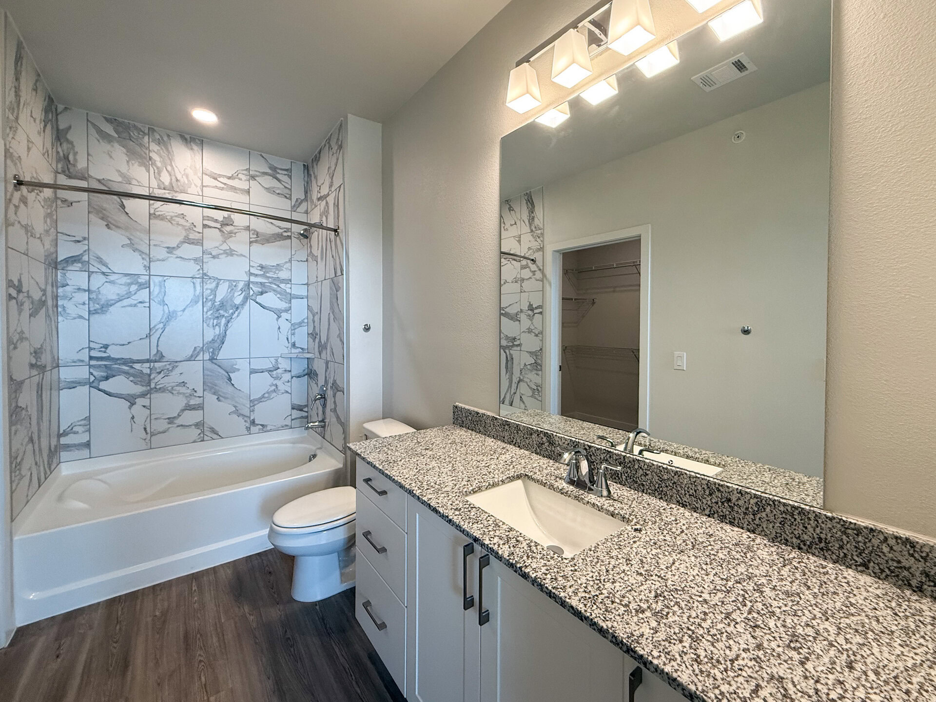 A modern bathroom features a white bathtub with a marble-patterned tile surround. The countertop is made of speckled gray granite, equipped with a rectangular sink and contemporary faucets. A large mirror above the sink reflects the space.