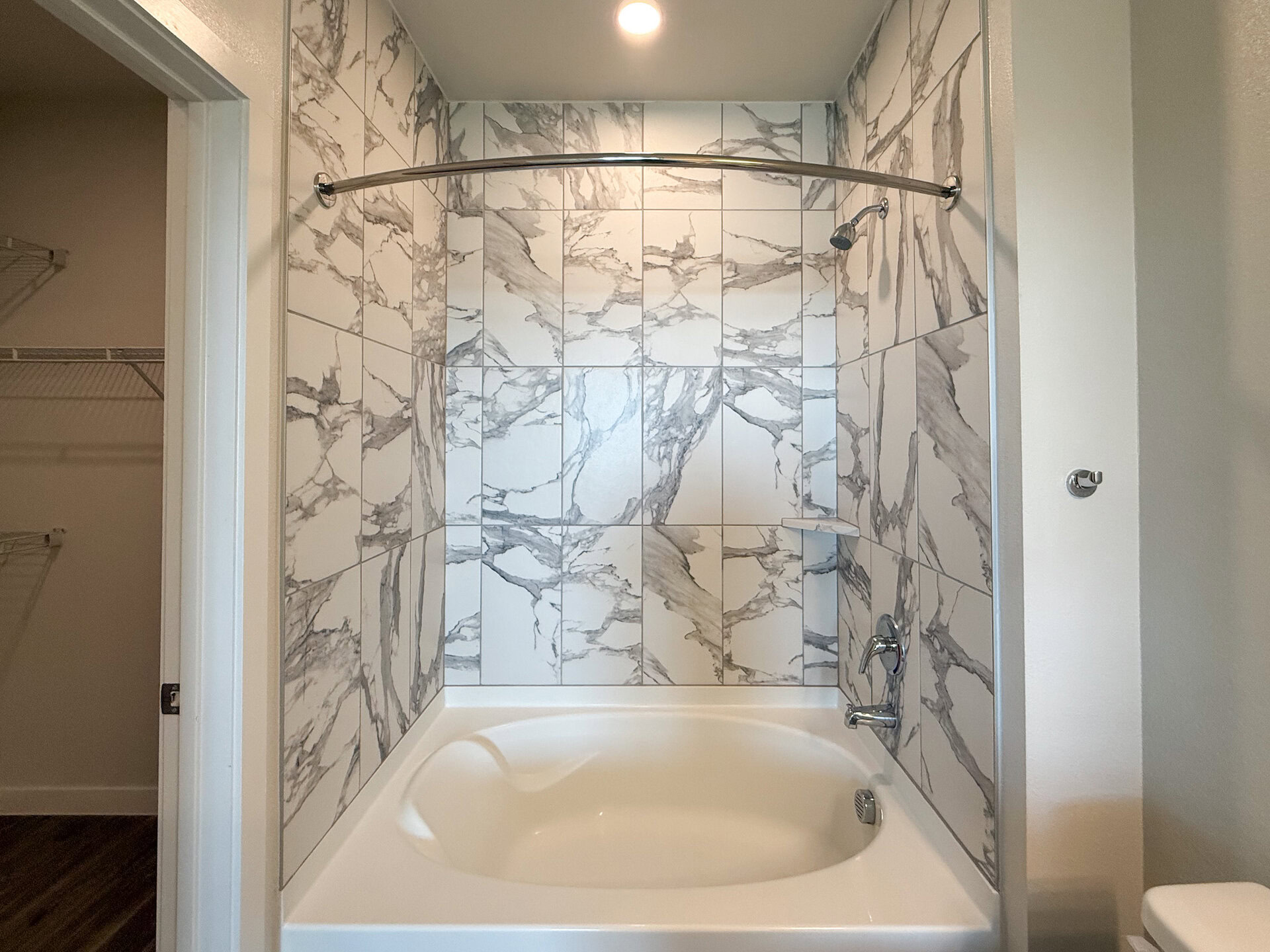 A modern bathroom features a bathtub with a curved shower rod above it. The walls are adorned with large, glossy tiles that have a marble-like pattern in shades of white and gray. A small shelf is integrated into the wall for convenience. The overall design is sleek and contemporary.