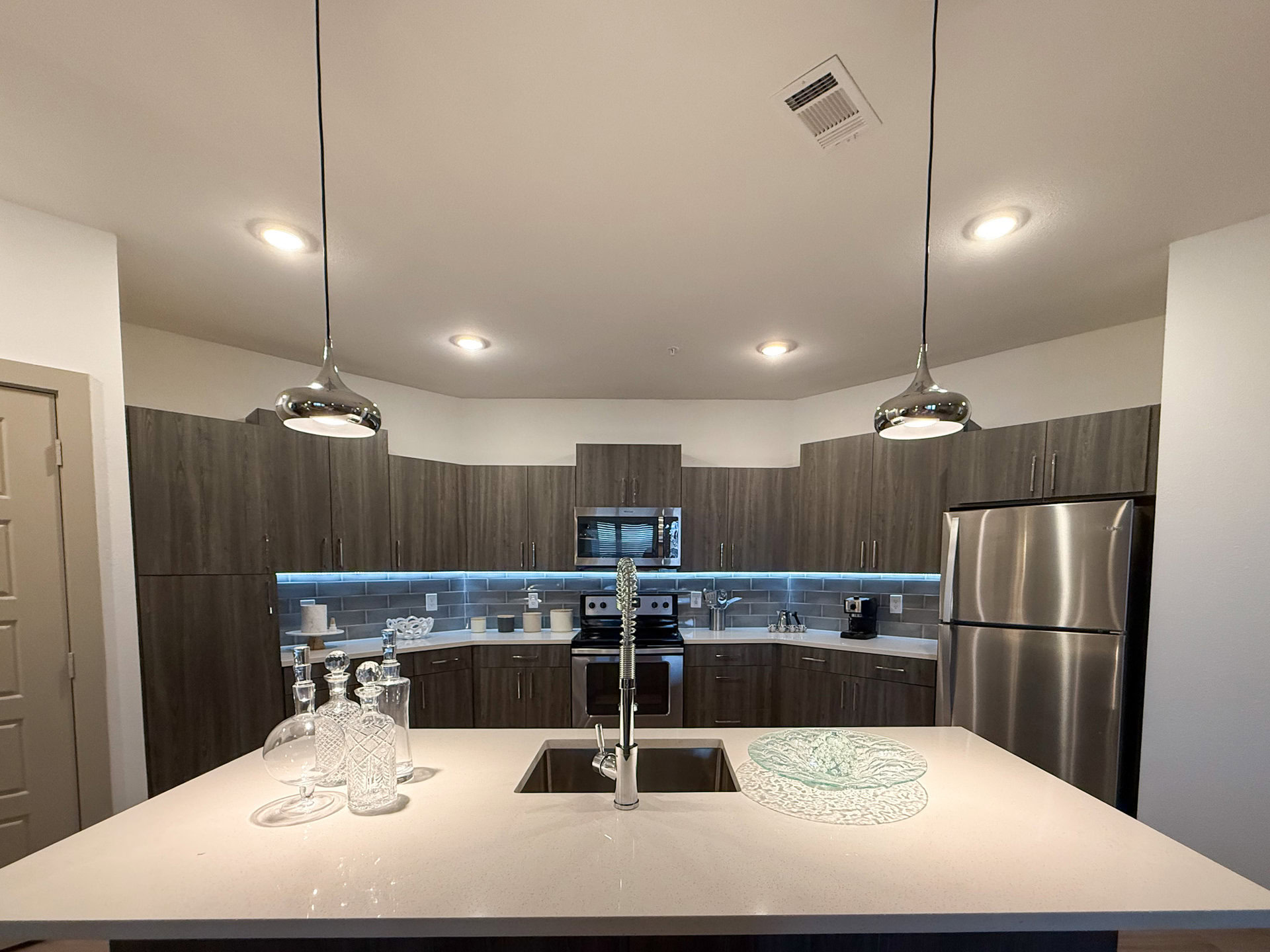 A modern kitchen featuring dark grey wood cabinetry and stainless steel appliances. The countertop is light-colored with a sleek design, and a stylish faucet is positioned over a square sink. Above the island, there are contemporary pendant lights. The backsplash is tiled in a subtle gray, and there is under-cabinet lighting that adds a warm glow. Beverage cooler is behind island,