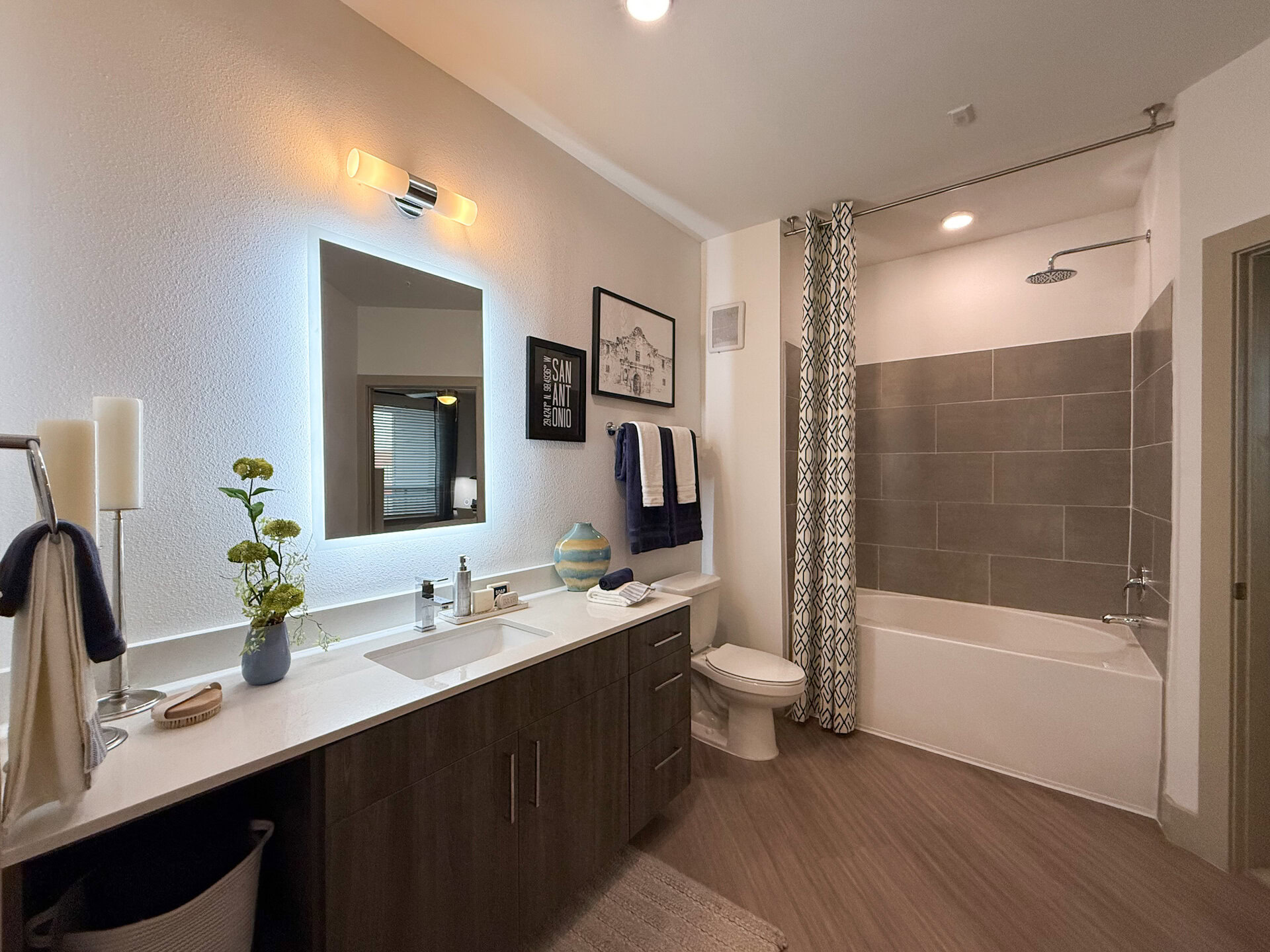 A modern bathroom features a double sink vanity with a white countertop and dark wood cabinetry. Above the sinks, a backlit mirror provides illumination.