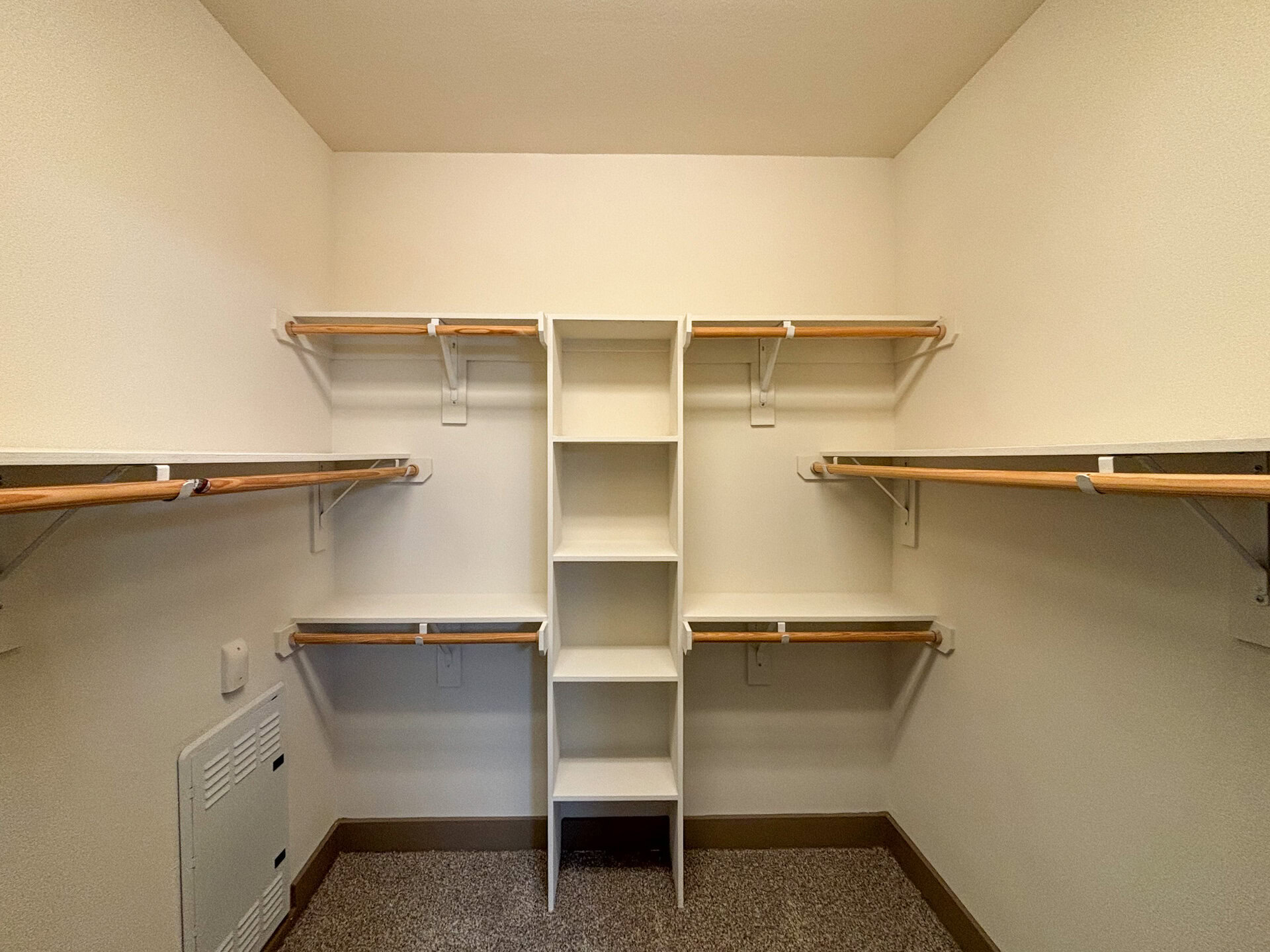 San Antonio Apartment's spacious closet features multiple wooden hanging rods on both sides, providing ample space for clothing. In the center, there is a tall, white shelving unit with five open compartments for storage.