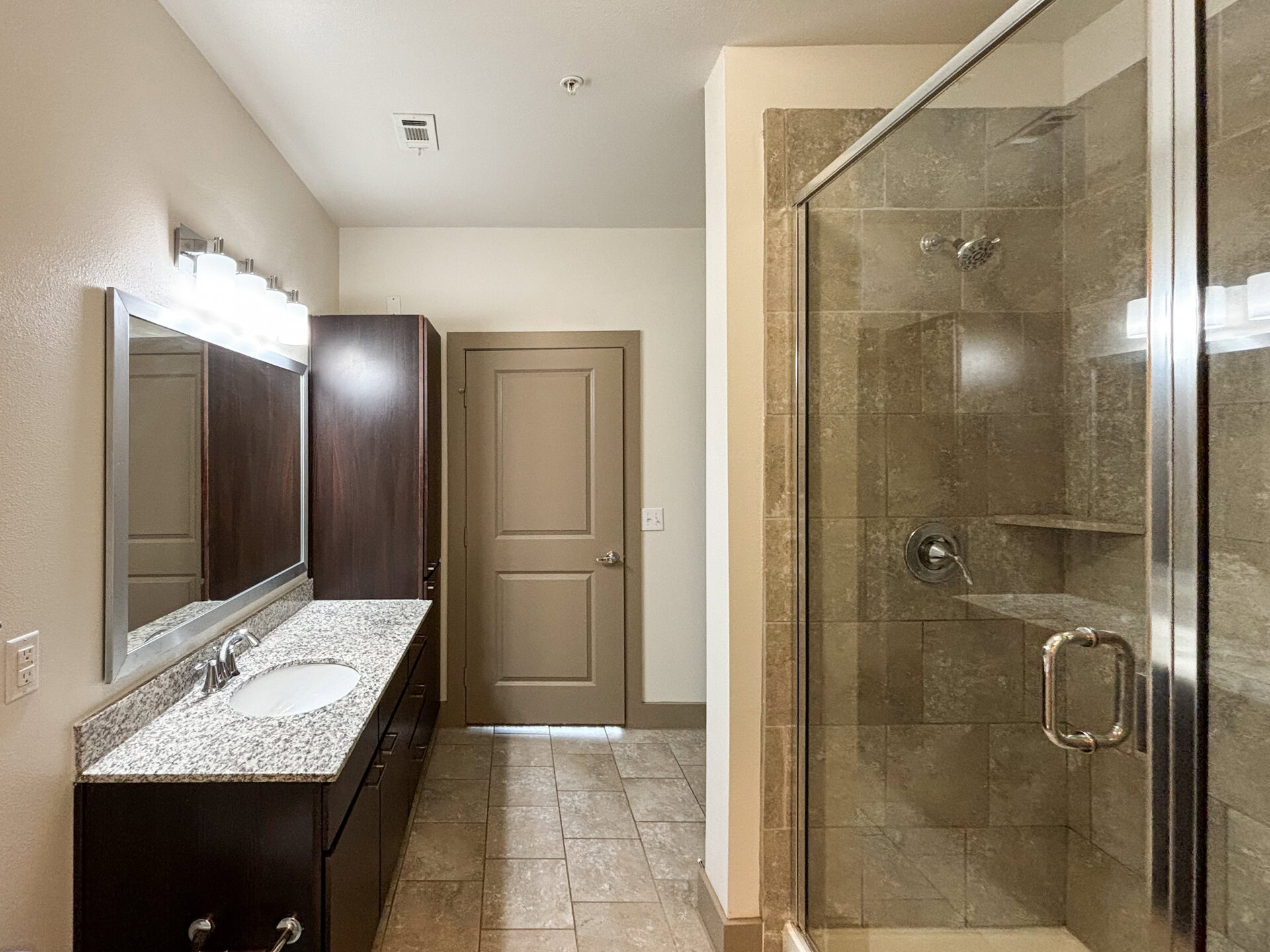 San Antonio apartment's modern bathroom features a sink vanity with a granite countertop and a large mirror above. The cabinetry is dark wood, providing a sleek contrast to the light-colored walls. To the right, a glass-enclosed standing shower showcases tiled walls and a built-in shelf.