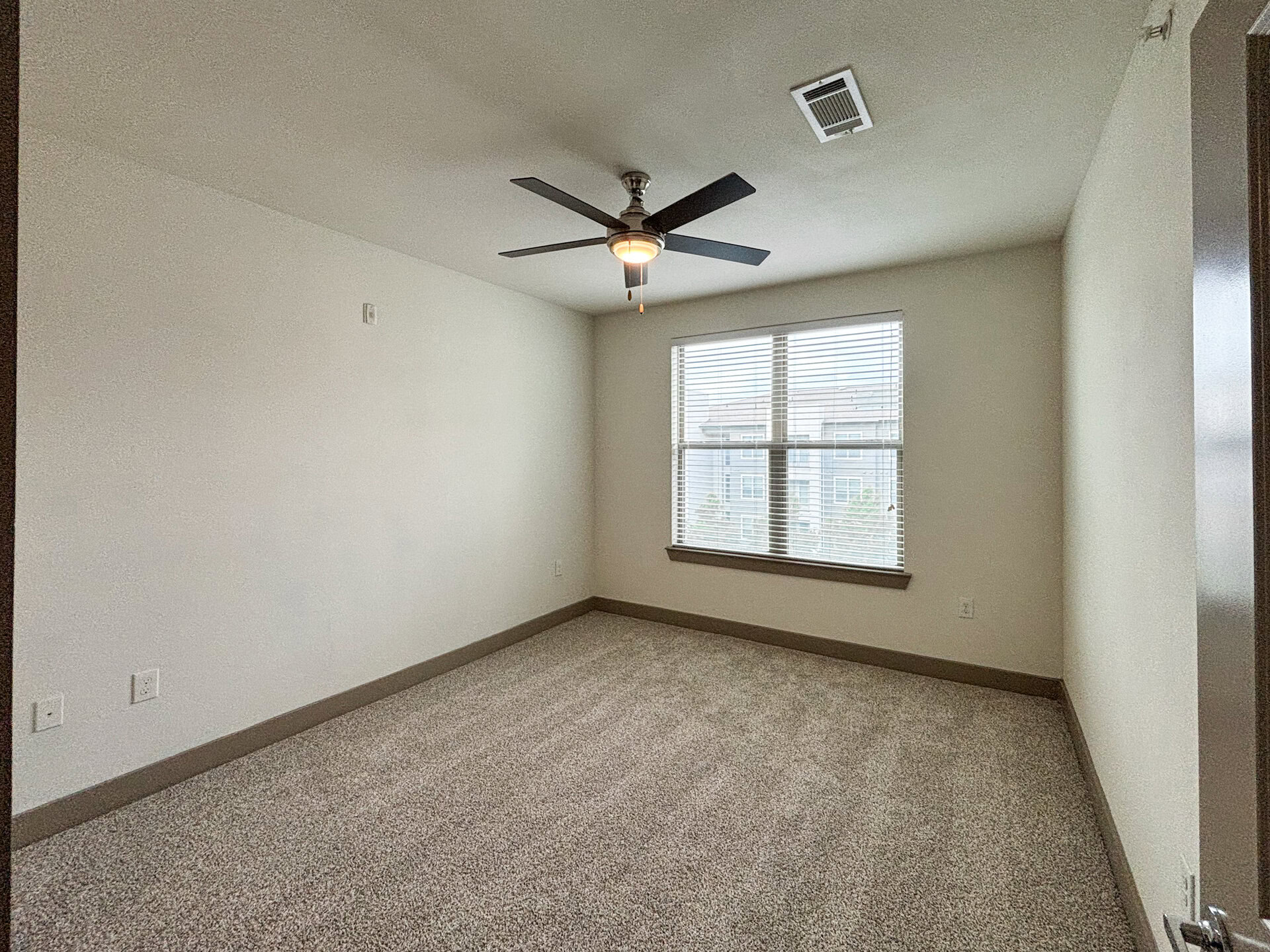 San Antonio apartment's spacious bedroom with light-colored walls and a plush carpeted floor. A ceiling fan with a light fixture hangs from the ceiling. There is a large window with horizontal blinds, allowing natural light to enter.