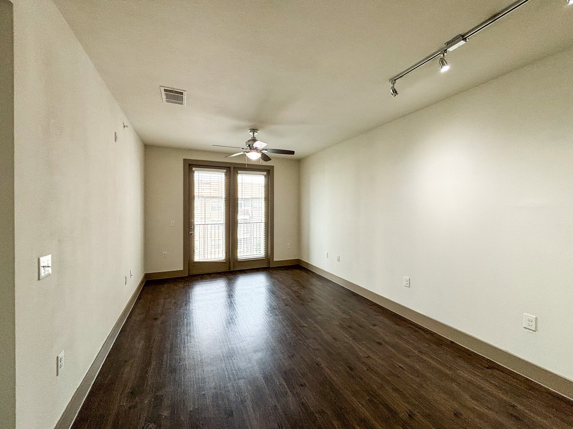 San Antonio's apartment spacious living room, empty room with light-colored walls and dark wood flooring. A ceiling fan with a light fixture is installed in the center. On one side, there are double doors with blinds, leading to a balcony. The overall ambiance is bright and airy.