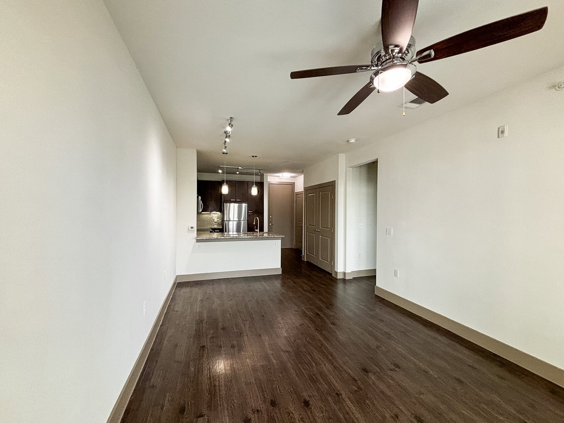 San Antonio apartment's spacious living area with dark wood flooring and light-colored walls. To the left, there is a kitchen featuring dark cabinetry, a stainless steel refrigerator, and a granite countertop with pendant lighting above. A ceiling fan with light is installed in the center of the room. The overall ambiance is modern and open.