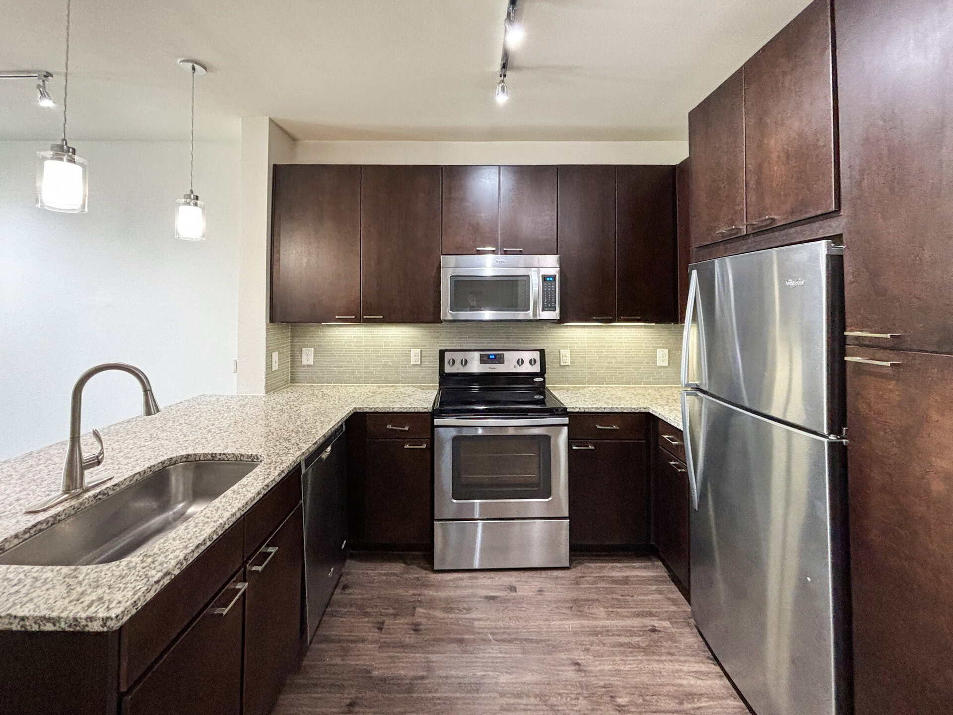 San Antonio apartment's modern kitchen featuring dark wood cabinetry and a light granite countertop. The stainless steel appliances include a microwave, oven, and refrigerator. A double-basin sink is integrated into the countertop, with a sleek faucet. The backsplash consists of light-colored tiles, and pendant lights hang above the counter, providing illumination. The flooring is a dark wood laminate, creating a warm contrast with the lighter elements.