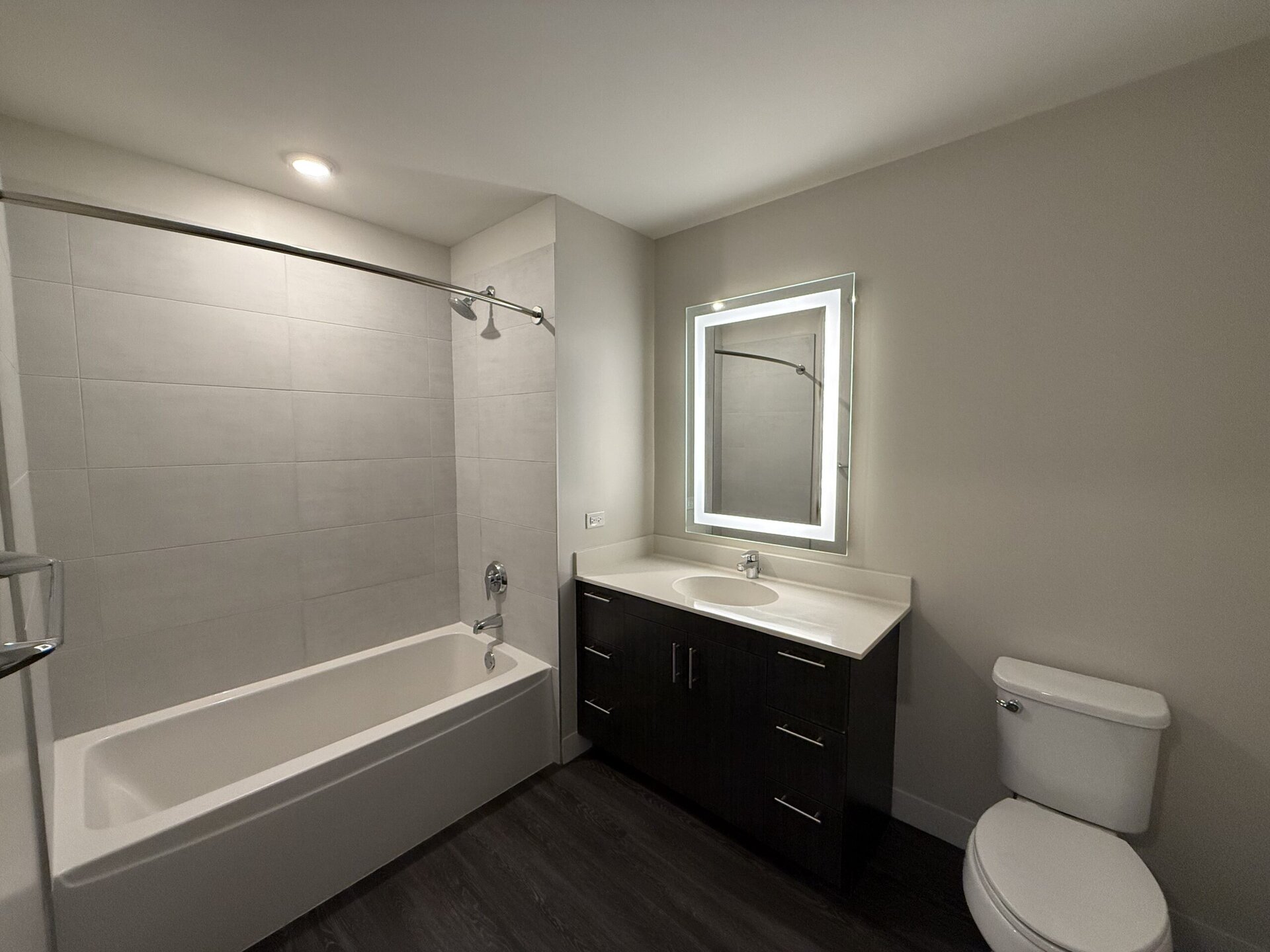 A modern bathroom featuring a bathtub with a showerhead above, surrounded by light gray tiles. To the right, there is a sleek vanity with a white countertop and a circular sink, complemented by dark wood cabinetry. Above the vanity, a rectangular mirror with built-in lighting is mounted on the wall. A white toilet is positioned next to the vanity, and the flooring is dark, adding contrast to the overall design. The space is well-lit with recessed ceiling lights.
