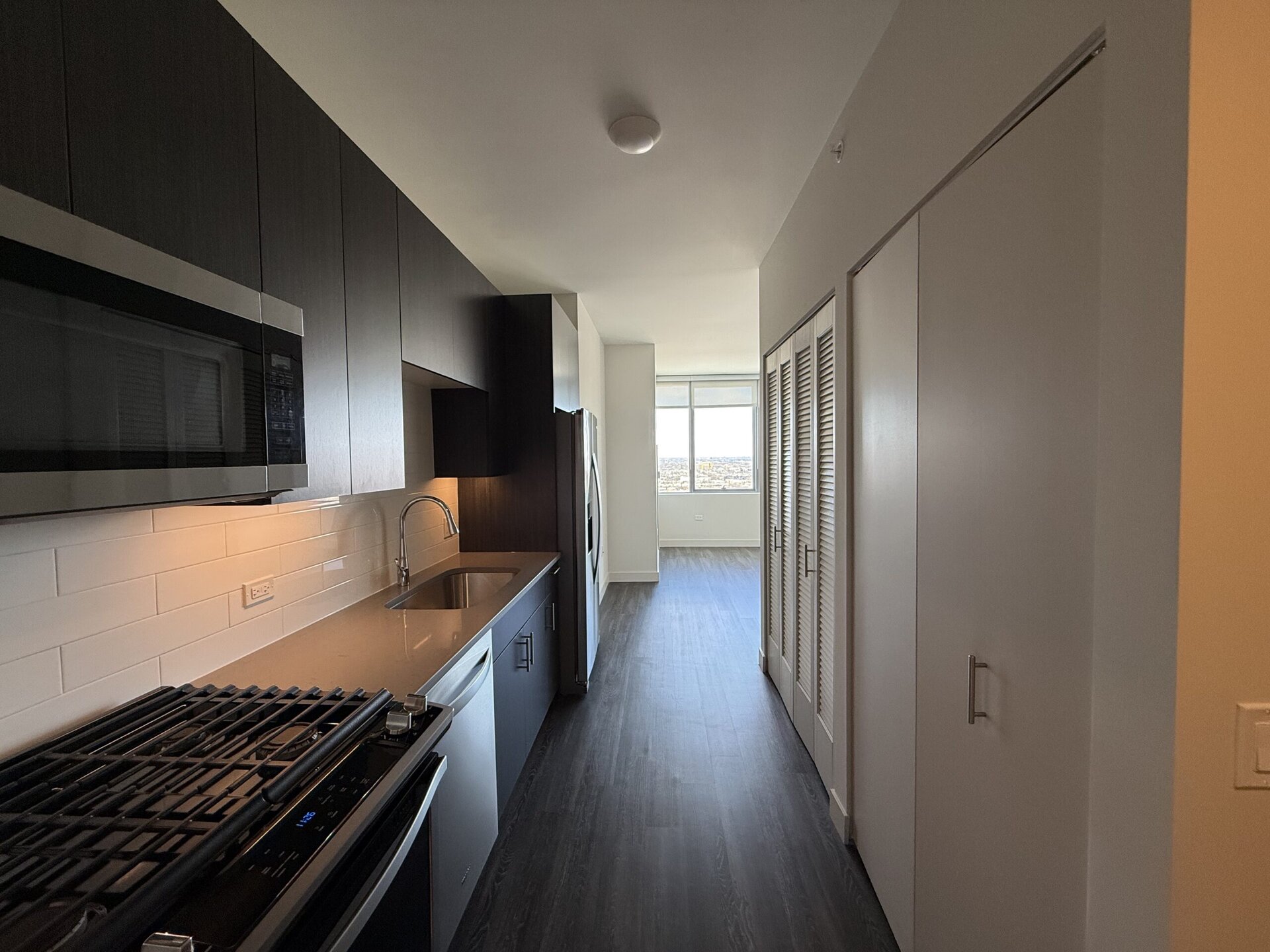 A modern kitchen area features dark cabinetry and a stainless steel microwave above a gas stove with a griddle. The countertop is a light color, complementing the white subway tile backsplash. To the right, a silver refrigerator stands next to a hallway leading to a bright room with large windows offering a view. On the right side of the hallway, there are double doors with louvered panels, likely for storage. The flooring is a dark wood laminate, creating a contemporary look.