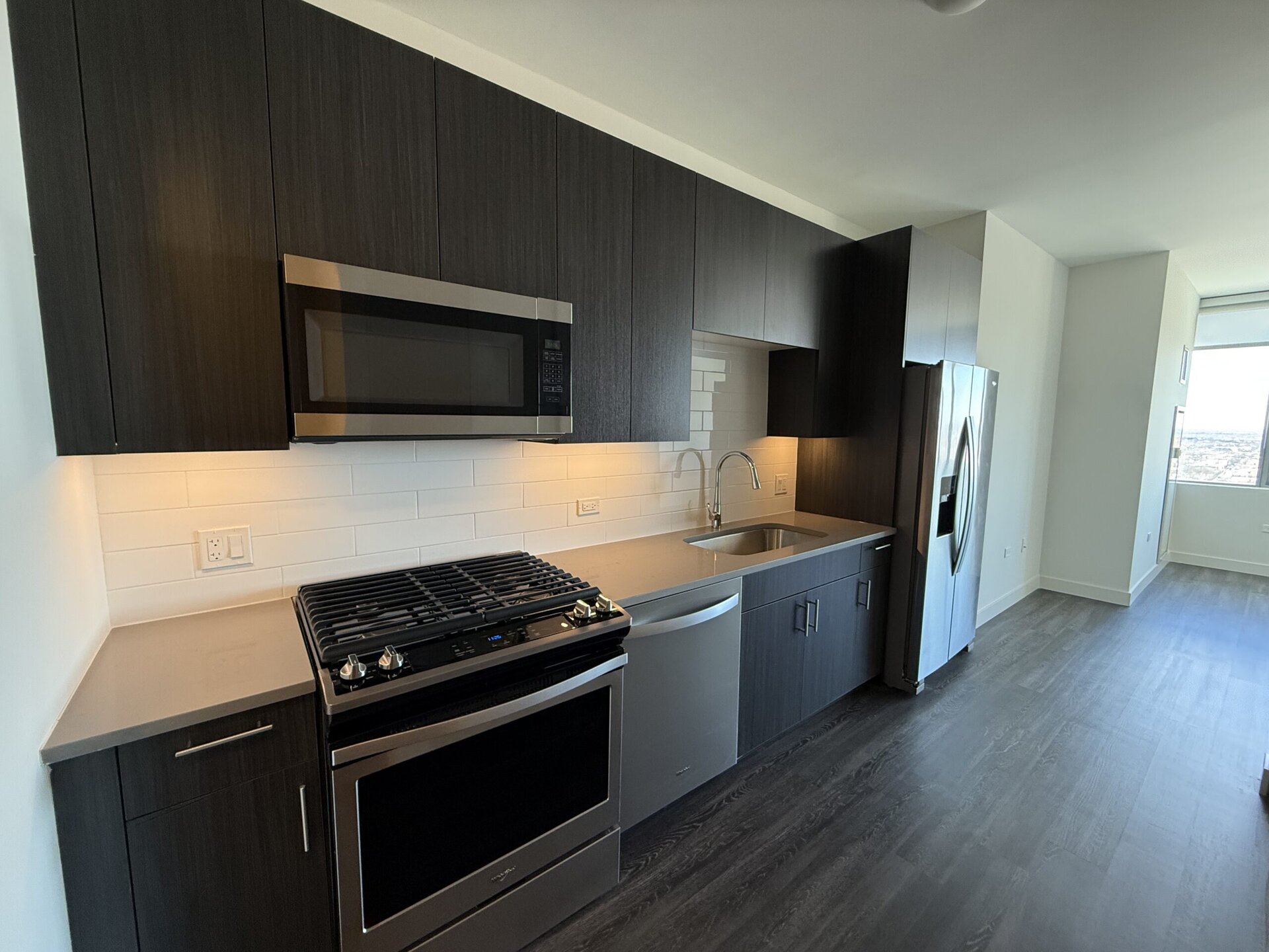 A modern kitchen features dark wood cabinetry and a light-colored countertop. The stainless steel appliances include a gas range with a griddle, a microwave mounted above, and a sleek refrigerator. A stainless steel sink is positioned under a window, with a contemporary faucet. The backsplash consists of white subway tiles, and the space is well-lit with under-cabinet lighting. The flooring is a light wood laminate, contributing to a clean and contemporary aesthetic.