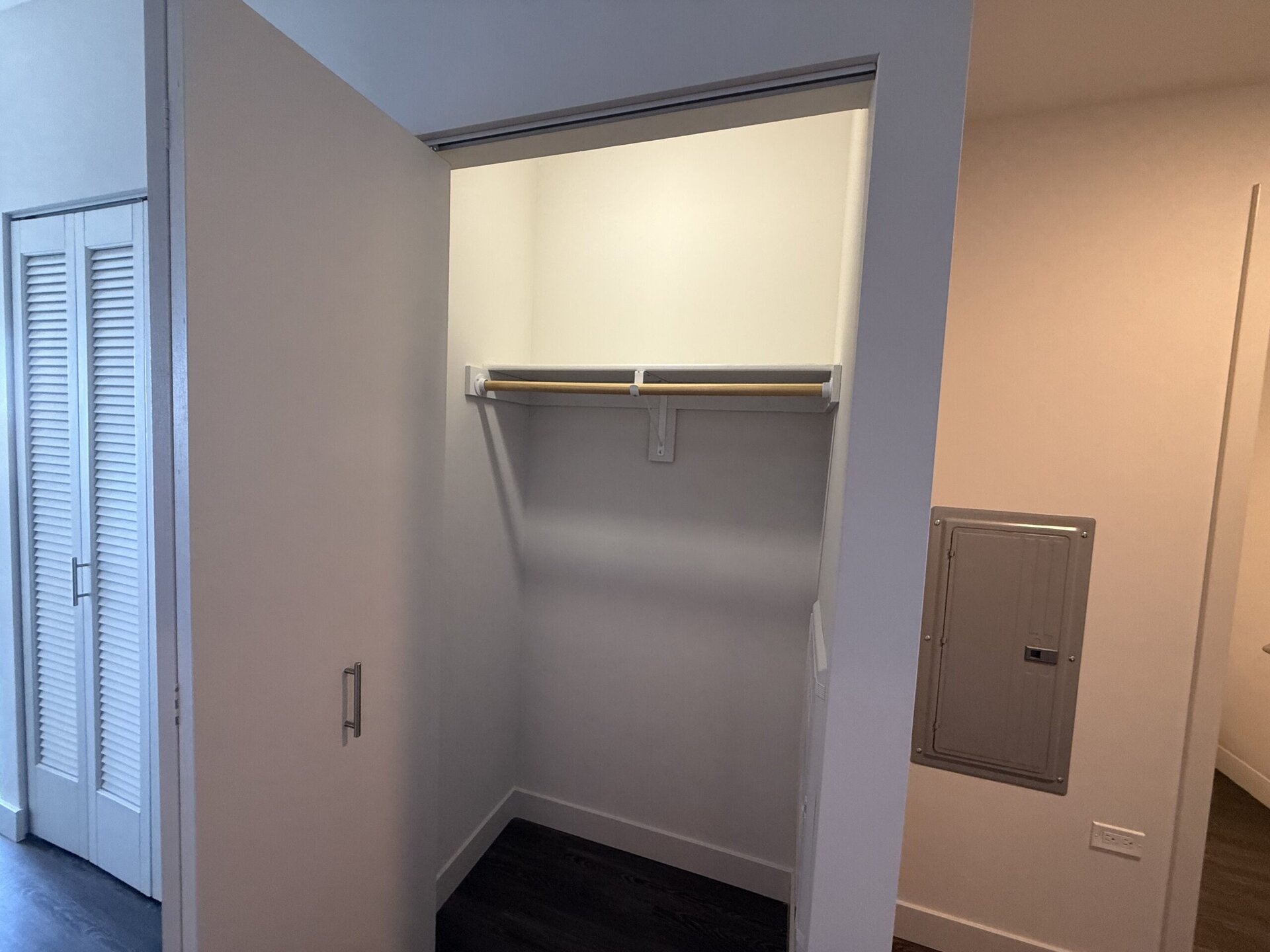 An empty closet is shown, featuring a single wooden hanging rod supported by white brackets. The walls are painted a light color, and the floor is dark. To the left, there is a set of louvered doors, likely leading to another storage space. On the right, a small electrical panel is mounted on the wall. The overall space appears clean and minimalistic.