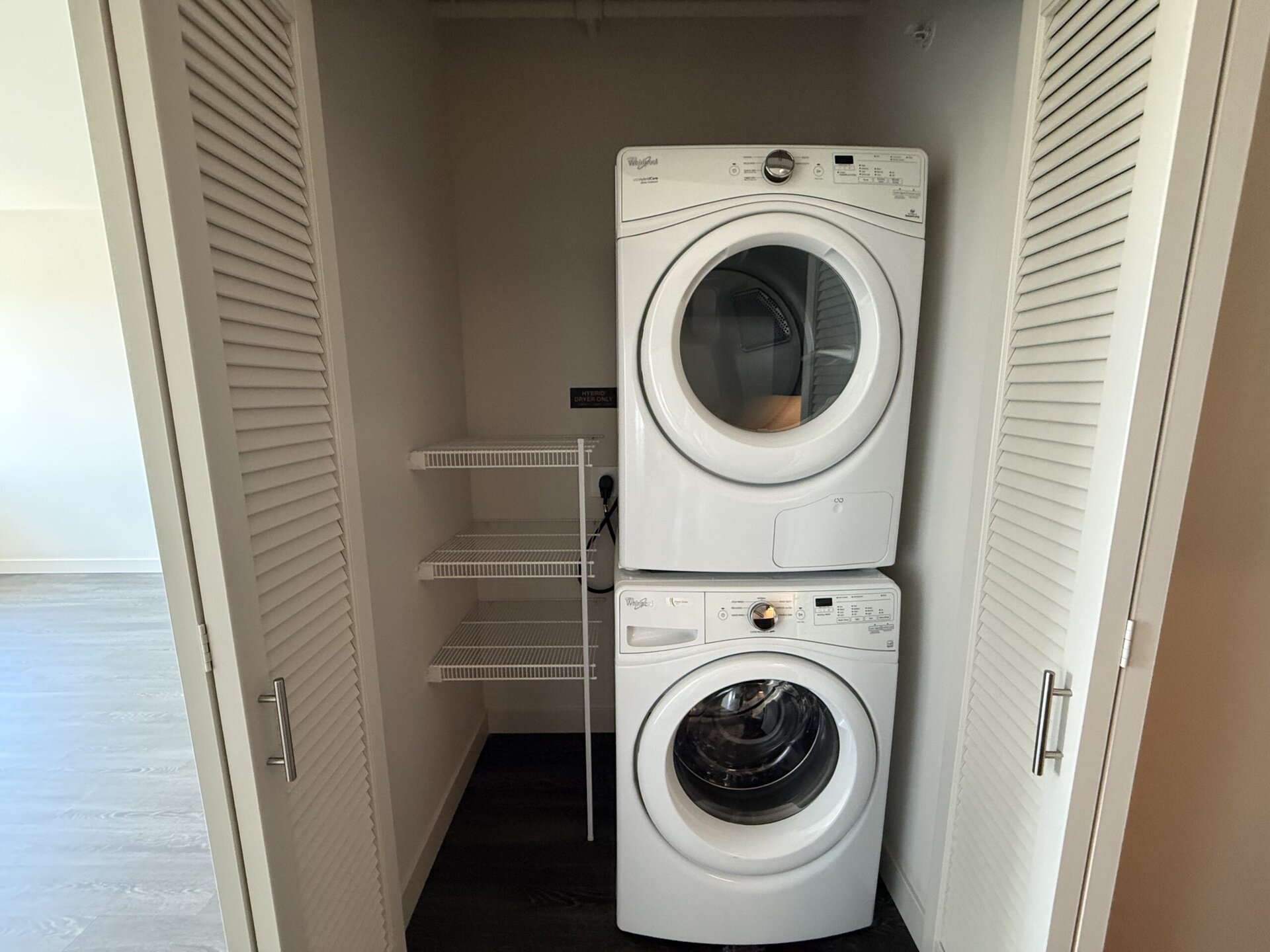 A stacked washer and dryer set is positioned in a closet space. The appliances are white, with the dryer on top and the washer below. To the side, there are two wire shelves for storage. The closet has a door with slatted panels, and the floor is a light-colored laminate. The background shows a neutral-colored wall and flooring.