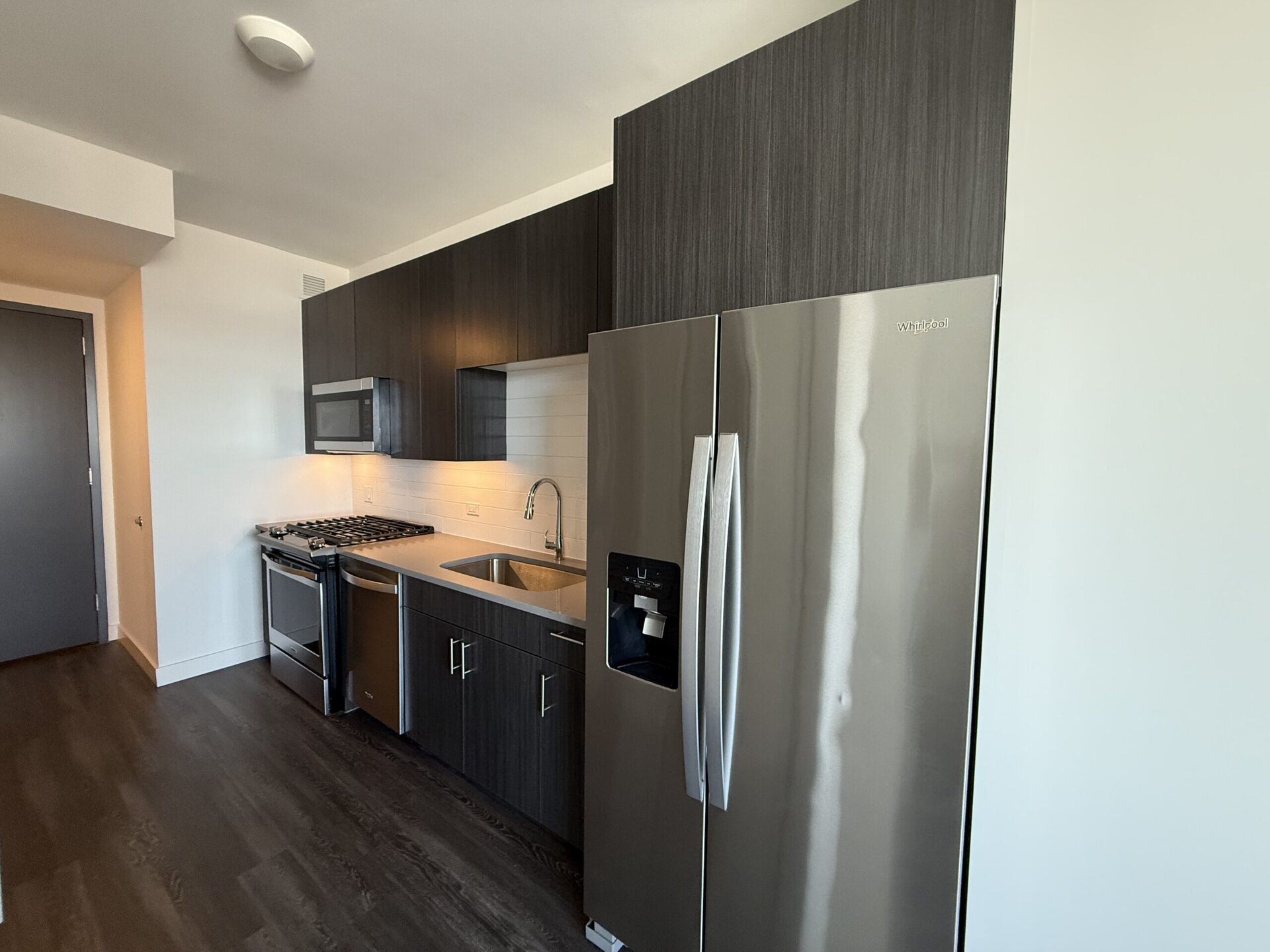 A modern kitchen features dark wood cabinetry and stainless steel appliances. The setup includes a gas stove, a microwave mounted above it, and a double-door refrigerator with a water dispenser. A stainless steel sink is positioned next to the stove, with a sleek faucet above it. The countertop is light-colored, contrasting with the dark cabinets, and the walls are painted white, enhancing the bright and contemporary feel of the space. The flooring is dark wood, adding warmth to the overall design.