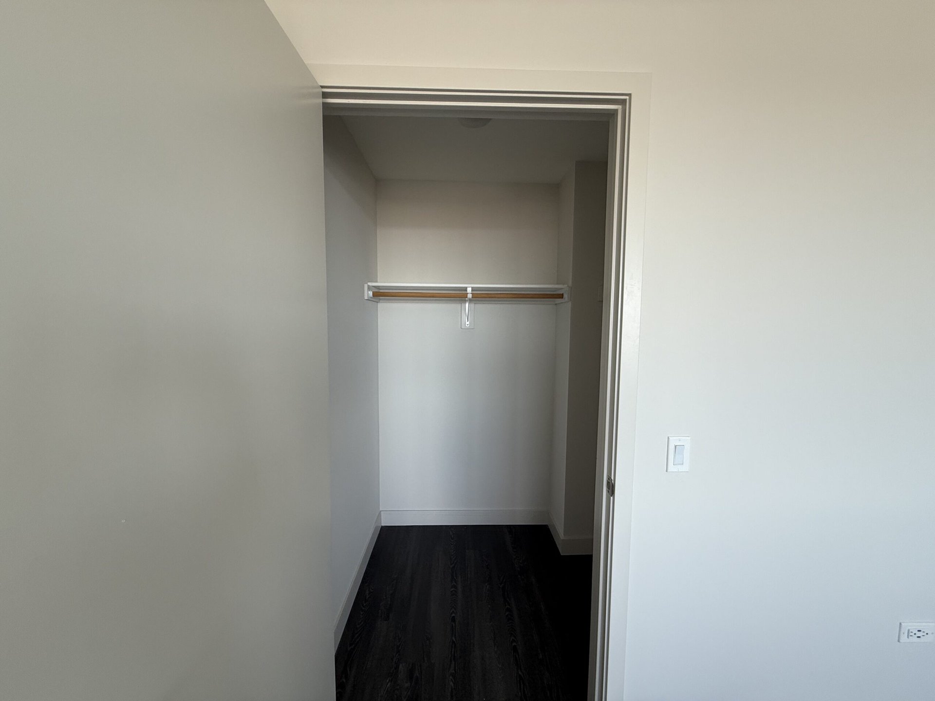A small, empty closet is visible, featuring a wooden hanging rod supported by metal brackets. The walls are painted a light color, and the floor is dark, likely hardwood. The closet has a simple, modern design with no shelves or additional storage features visible. A light switch is located on the right side of the entrance.