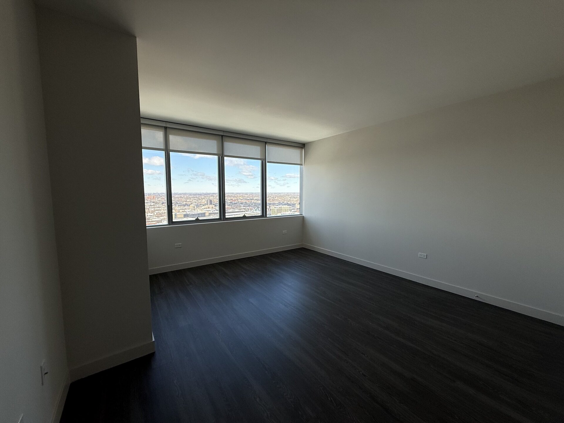 A spacious room with large windows offering a panoramic view of the city skyline. The walls are painted in a light color, and the flooring is dark, likely laminate or hardwood. The room appears empty, providing a sense of openness and potential for furnishing. Natural light floods in through the windows, enhancing the bright atmosphere.