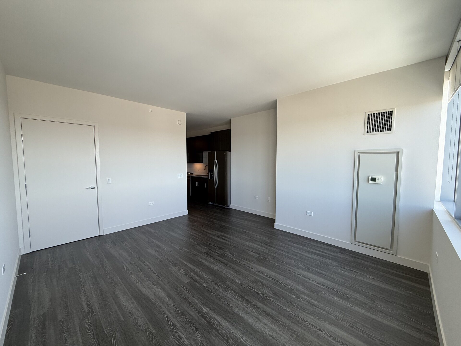 A spacious, modern room features light-colored walls and dark wood flooring. On the left, there is a closed door, while a small wall-mounted thermostat is visible on the right. In the background, a kitchen area with dark cabinetry and stainless steel appliances, including a refrigerator, is partially visible. A large window allows natural light to enter the space, enhancing the bright and airy atmosphere.