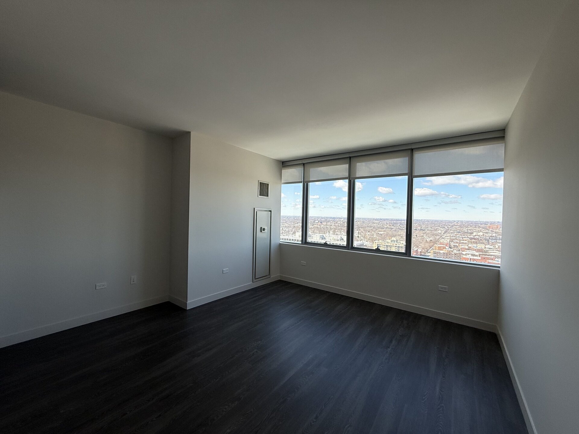 A spacious, empty room features large windows that offer a panoramic view of the cityscape below. The walls are painted in a light color, and the floor is dark wood laminate. A small closet is visible on one wall, and there is a vent and a thermostat mounted nearby. The overall atmosphere is bright and airy, with natural light flooding in from the windows.