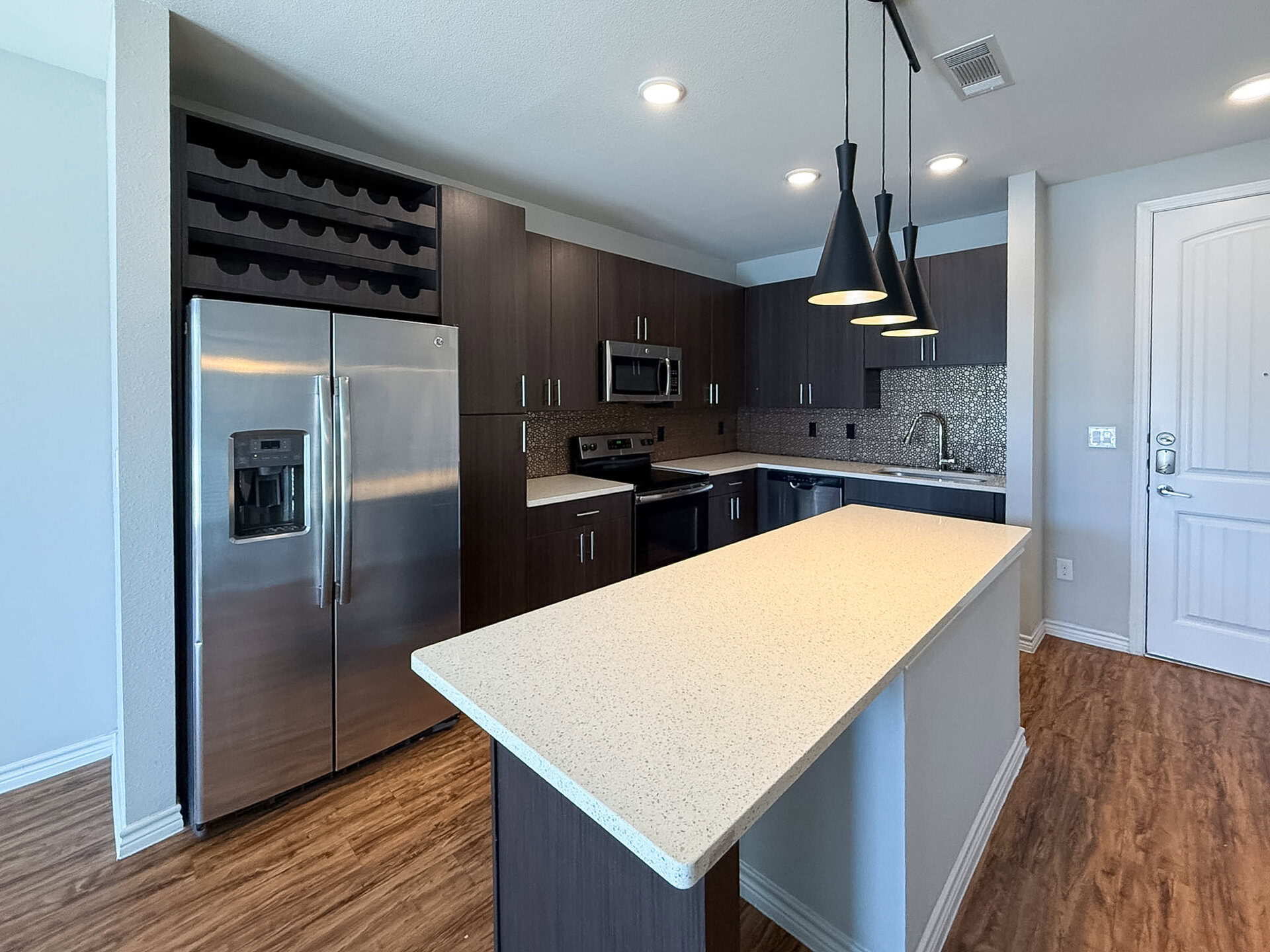 A modern kitchen featuring dark wood cabinetry and stainless steel appliances. The kitchen includes a large island with a light-colored countertop, providing ample workspace. Above the island, three black pendant lights hang down. The appliances include a side-by-side refrigerator and a wine rack. The backsplash is made of small, textured bronze tiles.