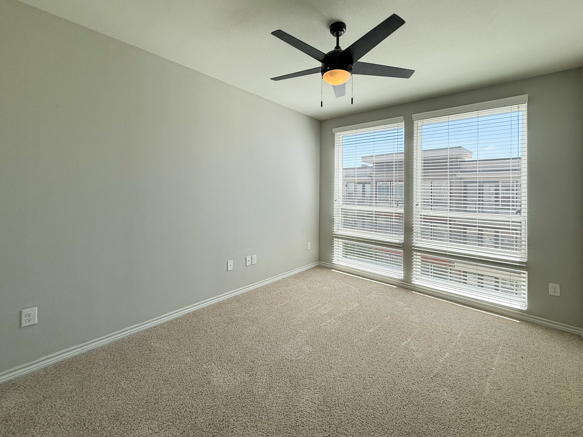 A well-lit bedroom features light gray walls and a plush carpeted floor. Two large floor-to-ceiling windows allow natural light to fill the space. A ceiling fan with black blades hangs from the ceiling, adding a modern touch.