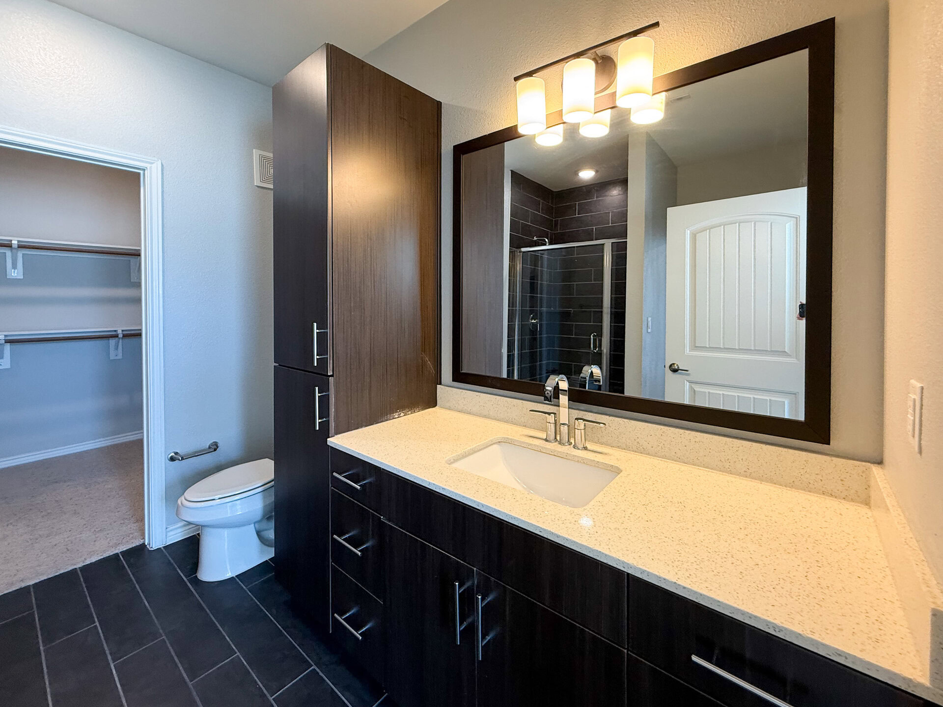 A modern bathroom features a sleek vanity with a white sink and polished chrome faucets, set against a light-colored countertop. The cabinetry is dark wood, providing a contemporary contrast. Above the sink, a large rectangular mirror is framed in dark wood. To the side, a glass standing shower enclosure with black tiles is visible.