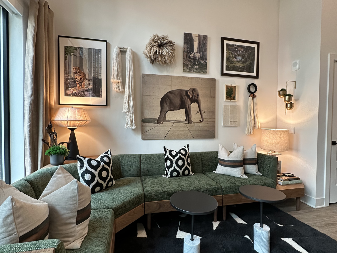 A cozy living room features a green sectional sofa adorned with various patterned cushions, including black and white designs. On the wall behind, a collection of framed artwork includes images of animals and abstract pieces. A stylish lamp with a textured shade sits beside the sofa, illuminating the space. Two small black side tables with marble bases are positioned in front of the sofa, and a dark area rug adds warmth to the floor. Natural light streams in through large windows, enhancing the inviting atmosphere.