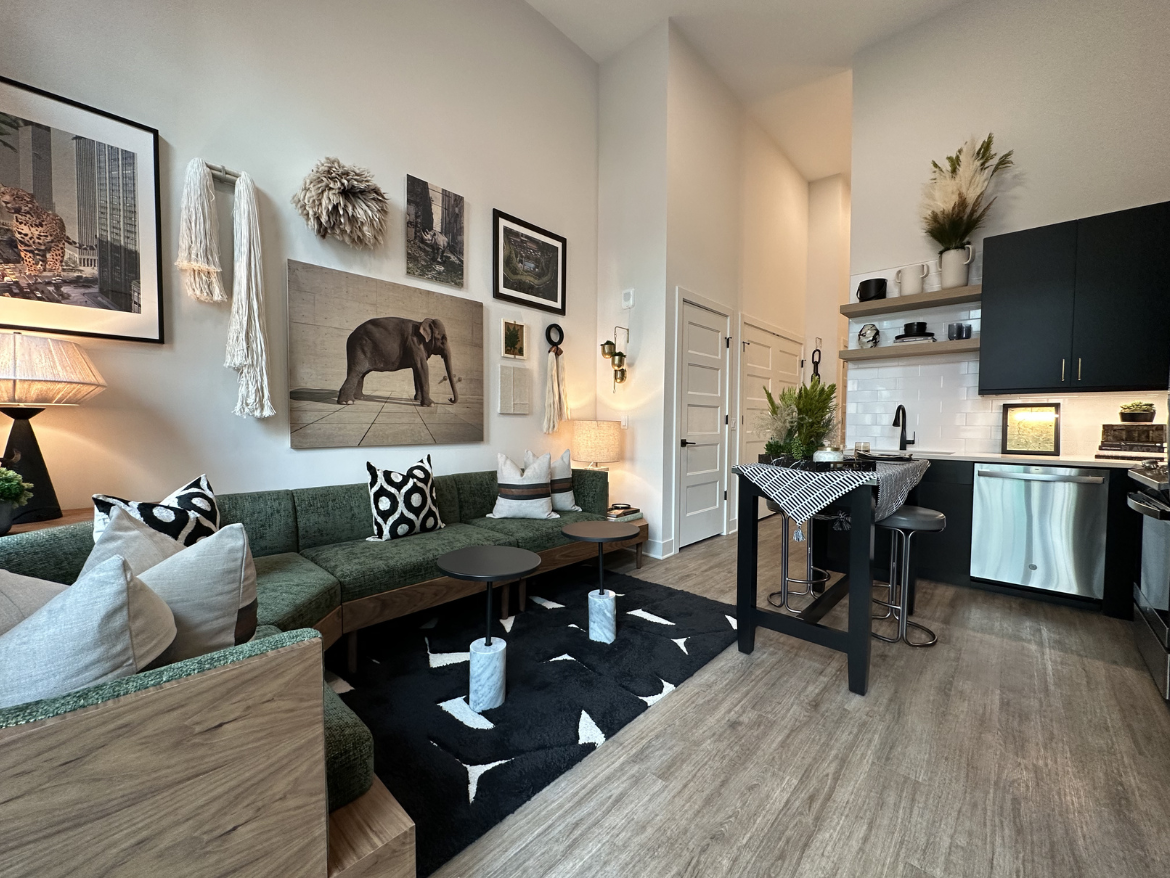 A modern living space features a green sectional sofa adorned with various patterned pillows. The wall behind is decorated with framed artwork, including a prominent image of an elephant. A cozy lamp stands next to the sofa, illuminating the area. In the kitchen, sleek black cabinetry contrasts with white tiles, and a small dining table with a checkered tablecloth is set with decorative plants. The flooring is a light wood, enhancing the contemporary feel of the room.