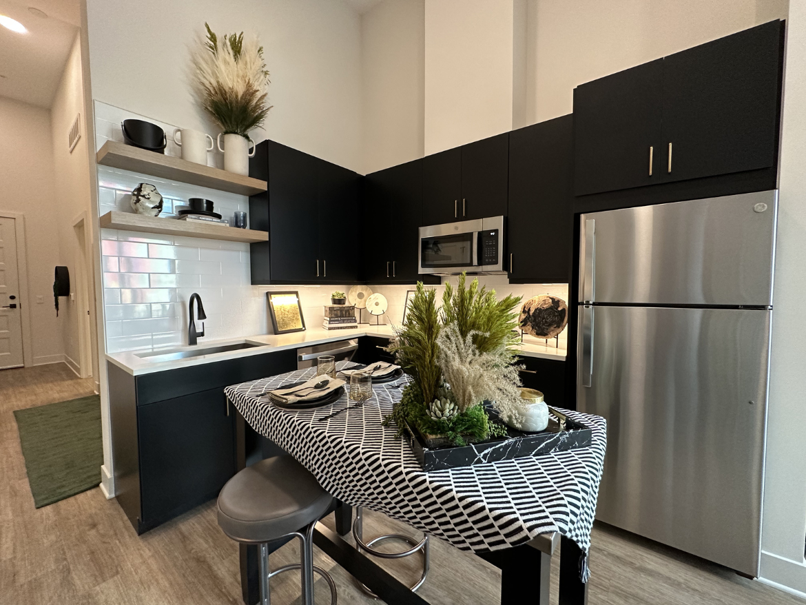 A modern kitchen features sleek black cabinetry and stainless steel appliances, including a microwave and refrigerator. A white countertop complements the cabinetry, with a stylish black and white tablecloth draped over a central island. The island is adorned with a decorative arrangement of greenery and small decorative items. Above the countertop, open shelves display various kitchenware and decorative pieces. The space is well-lit, with a light-colored backsplash and a warm wooden floor, creating a contemporary and inviting atmosphere.