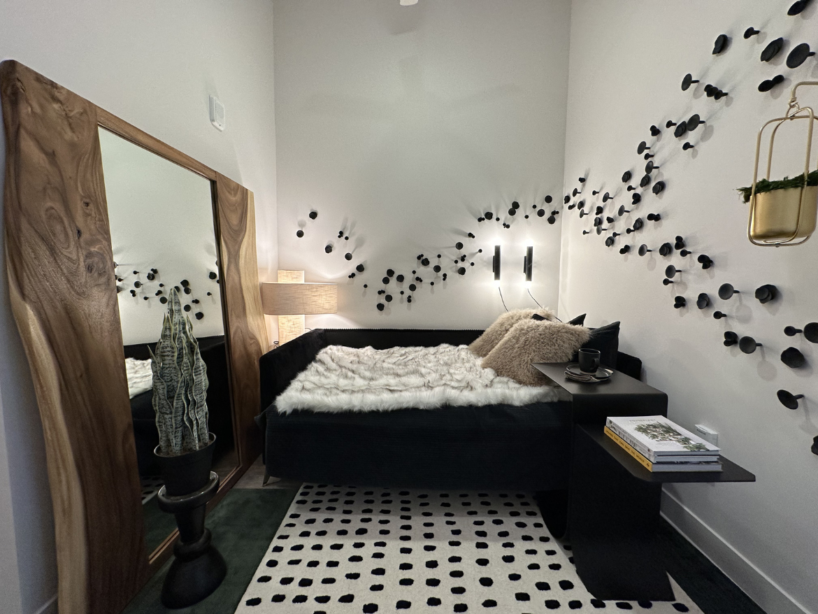 A cozy bedroom features a black bed with a fluffy white throw and decorative pillows. A large, natural wood-framed mirror leans against the wall beside the bed. The wall is adorned with a unique arrangement of black circular wall decor. A stylish lamp provides soft lighting, and a small side table holds a book and a cup. A potted plant adds a touch of greenery, and a patterned rug with black dots complements the space.