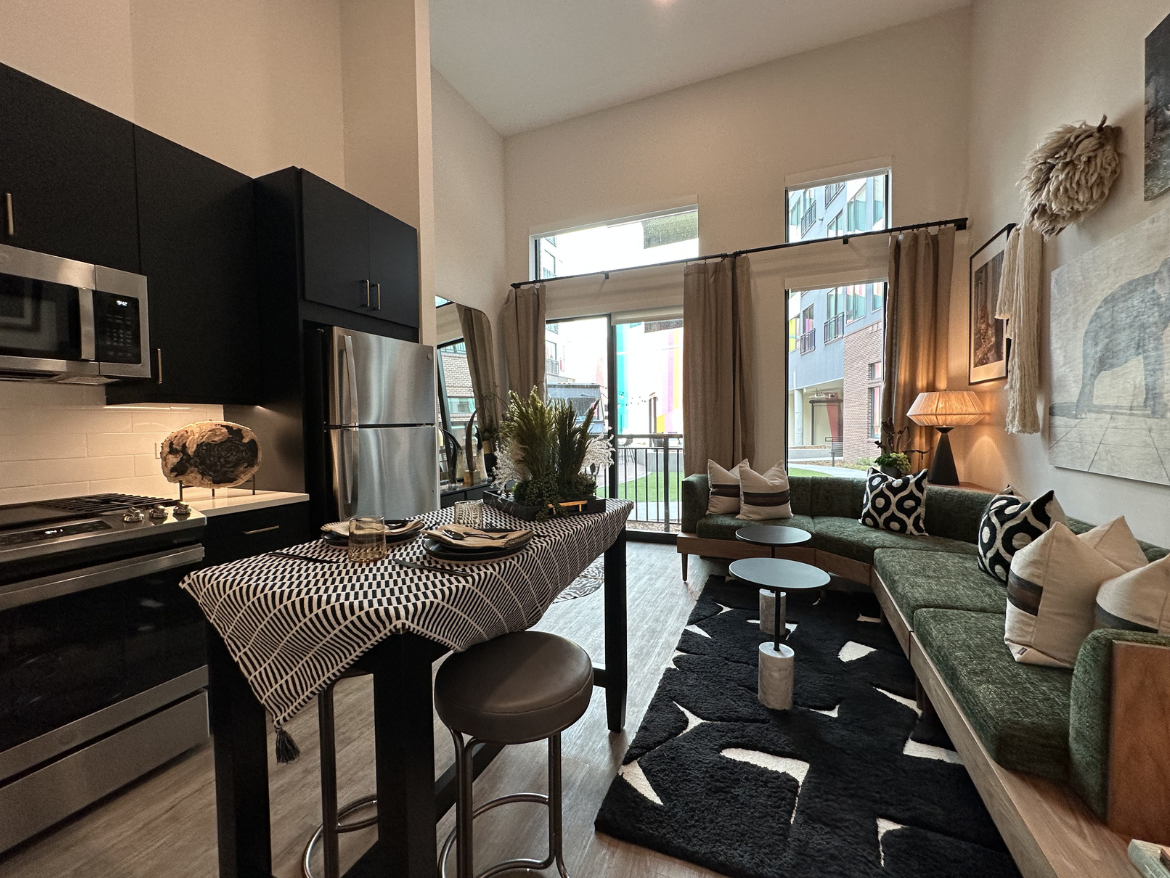 A modern, stylish living space featuring an open kitchen and living area. The kitchen has sleek black cabinetry, stainless steel appliances, and a countertop with a decorative stone piece. A dining area with a patterned table runner is set with plates and glasses. The living area includes a green sectional sofa adorned with patterned cushions, a couple of small round tables, and a textured black rug. Large windows allow natural light to fill the room, with sheer curtains framing the view of an outdoor space. Decorative elements like wall art and a unique lamp add character to the space.