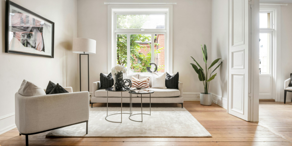 A bright and airy living room of a short-term lease apartment features a light-colored sofa adorned with black and white cushions, complemented by a small coffee table with a circular design. A floor lamp stands beside the sofa, and a large window allows natural light to flood the space, revealing greenery outside. A potted plant adds a touch of nature, while a soft area rug covers the wooden floor, enhancing the cozy atmosphere. The walls are painted in a neutral tone, and a framed artwork hangs on one side, contributing to the room's modern aesthetic.