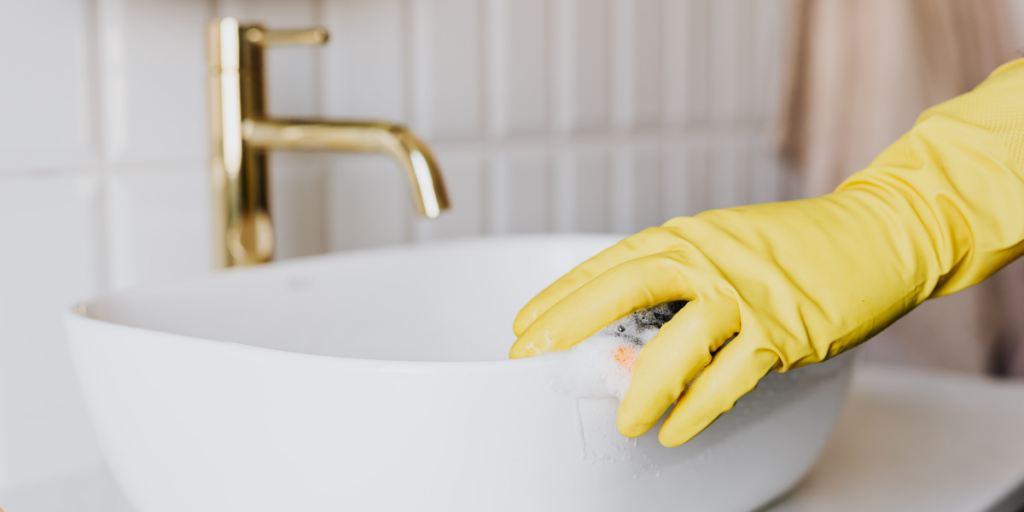 A hand wearing a yellow rubber glove is scrubbing the edge of a white, oval sink with a sponge that has soap suds on it. In the background, a gold faucet is visible, along with white tiled walls. The scene conveys a sense of cleanliness and home maintenance through spring cleaning