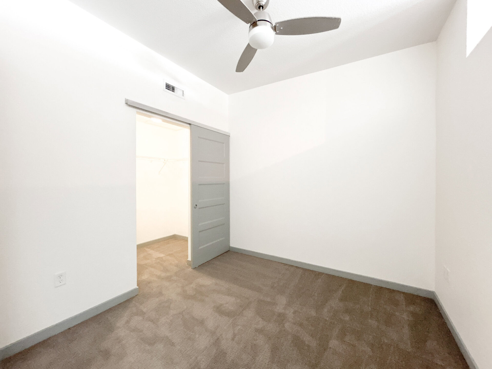 A small, empty room with light-colored walls and a light brown carpet. There is a gray door partially open, leading to a closet with a hanging rod visible inside. A ceiling fan is mounted above, and natural light enters through a small window. The overall atmosphere is bright and minimalistic.