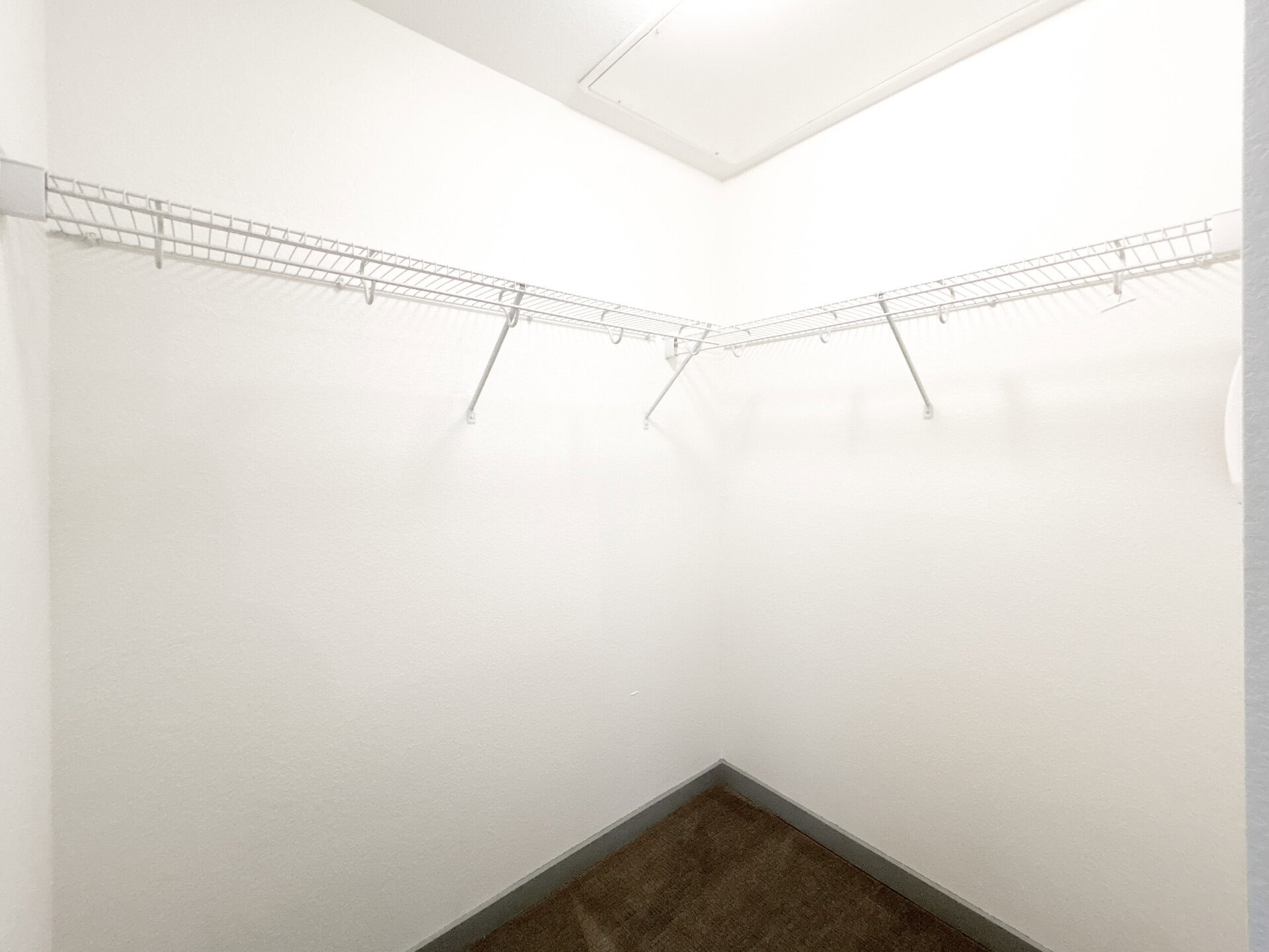 A small, empty closet features two wire shelving units that extend along the walls, forming a corner. The walls are painted white, and the floor is carpeted in a light brown color. The overall space is bright and uncluttered, emphasizing the simplicity of the design.