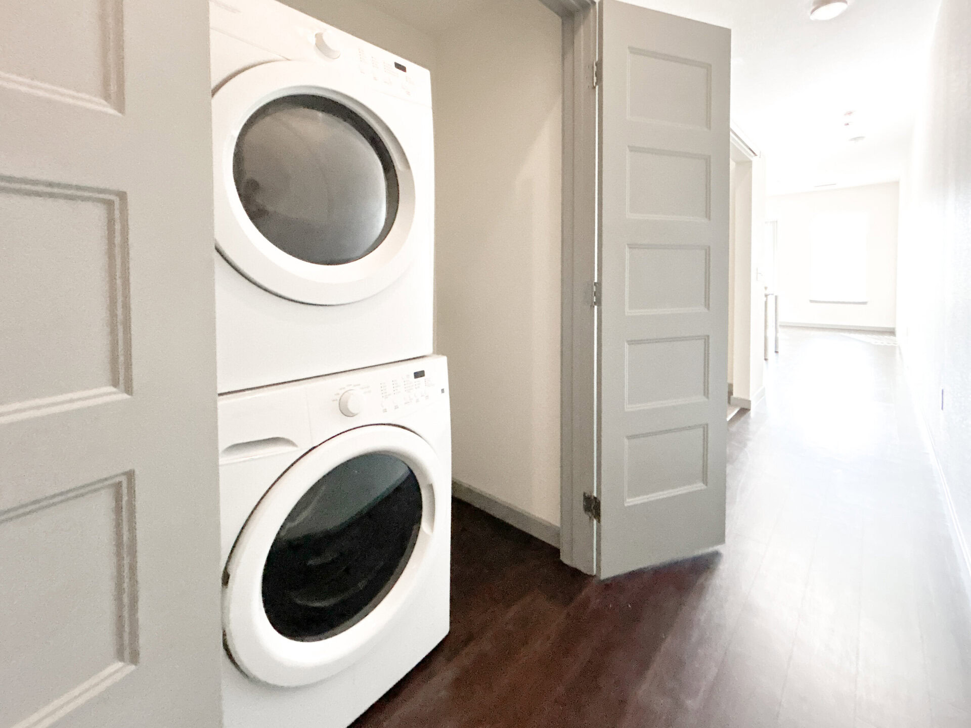 A stacked washer and dryer unit is positioned against a wall in a narrow hallway. The appliances are white, with a front-loading design. A partially open door is visible on the left, leading to another room. The hallway features dark wood flooring and a bright area at the far end, suggesting natural light from a window. The walls are painted in a light color, enhancing the spacious feel of the area.
