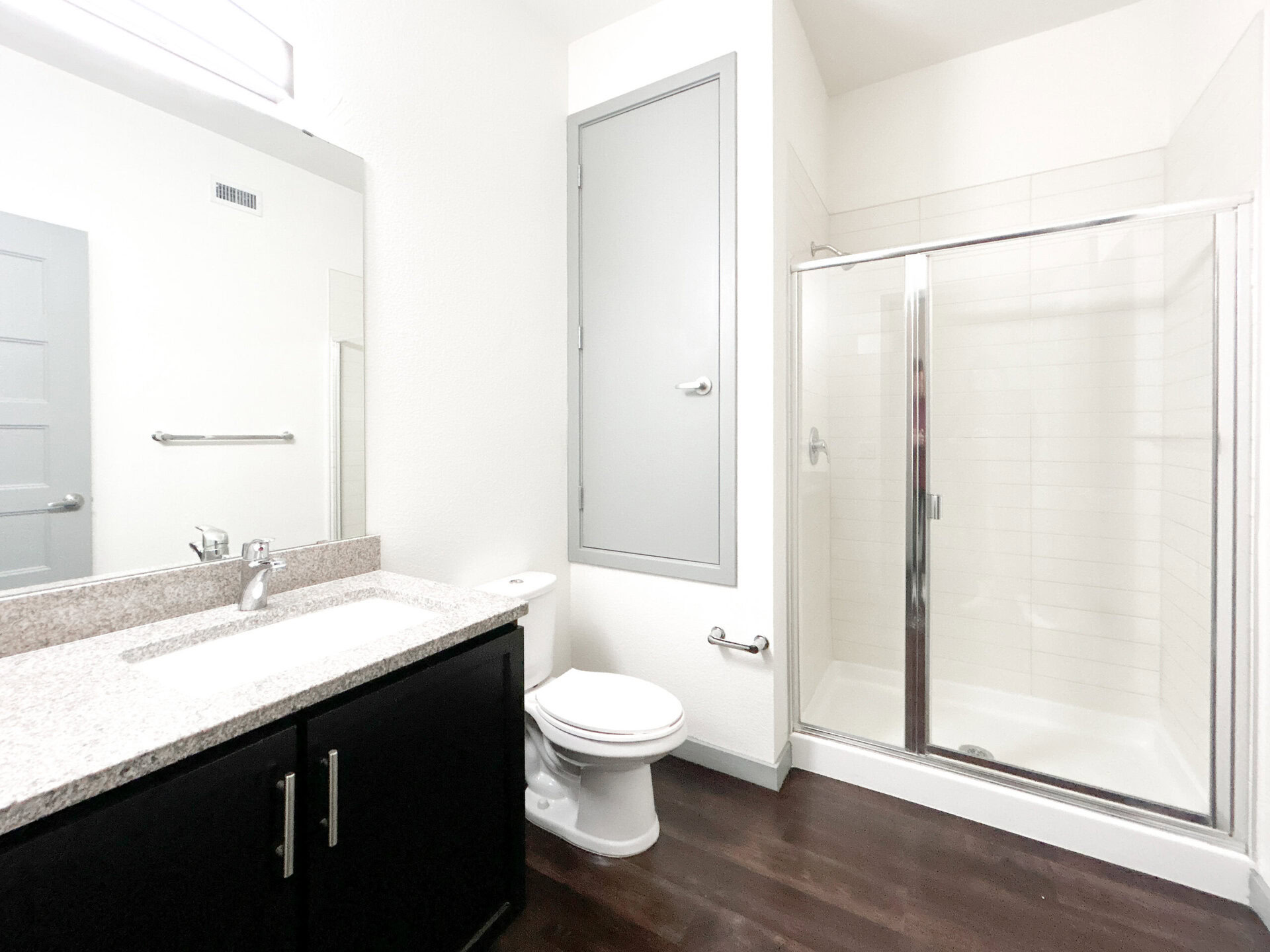 A modern bathroom featuring a sleek design. The space includes a granite countertop with an undermount sink and a polished chrome faucet. Below the countertop, there are dark cabinetry doors. To the left, a white toilet is positioned next to a gray door leading to a storage area. A glass shower enclosure with sliding doors is visible, showcasing white tiled walls and a simple showerhead. The flooring is dark, contrasting with the light-colored walls and fixtures. A large mirror above the sink reflects the room's bright ambiance.