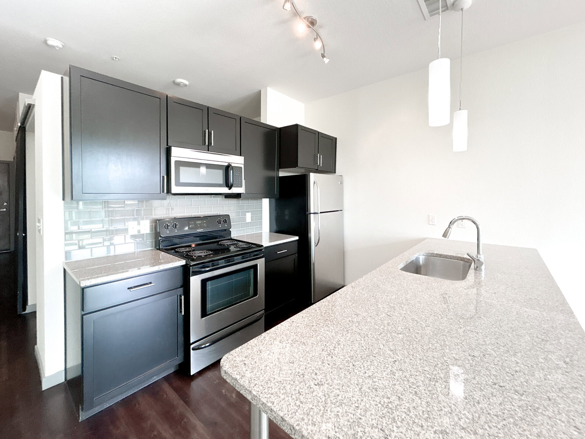 A modern kitchen featuring dark cabinetry and stainless steel appliances. The kitchen includes a gas stove, microwave, and a refrigerator. A light-colored granite countertop extends into a small island with a sink. The backsplash consists of light gray tiles, and there are two pendant lights hanging above the island. The flooring is dark wood, creating a contrast with the lighter elements in the kitchen.