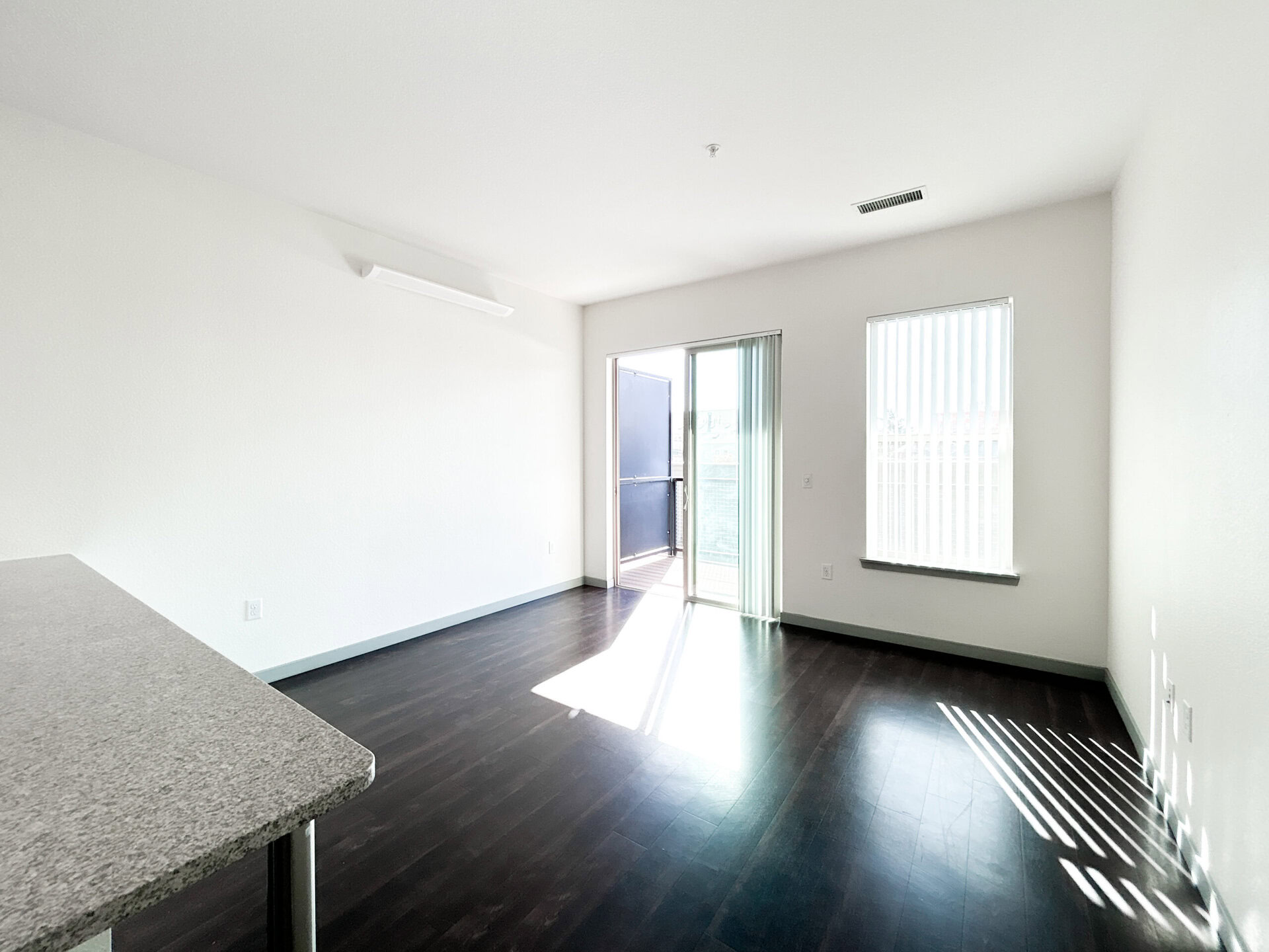 A bright, spacious room features dark hardwood flooring and white walls. On one side, there is a sliding glass door leading to a small balcony, allowing natural light to fill the space. Adjacent to the door, a window with vertical blinds provides additional light. A countertop is visible in the foreground, suggesting a kitchen area. The overall atmosphere is modern and inviting.