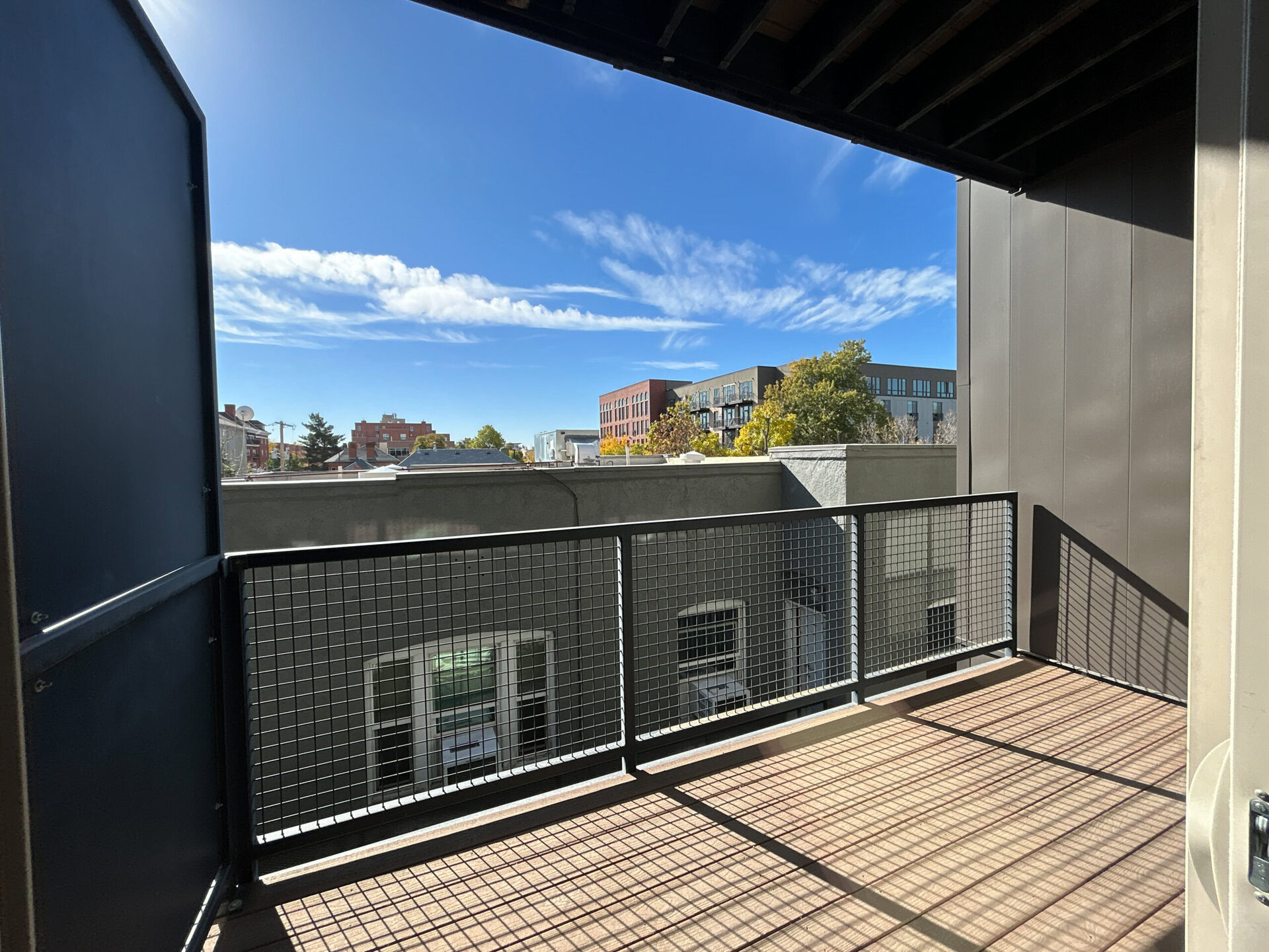 A balcony view showcases a clear blue sky with wispy clouds. The foreground features a black metal railing with a grid pattern. Below, rooftops of nearby buildings are visible, including a mix of residential and commercial structures, some with greenery. The scene is bright and sunny, indicating a pleasant day.