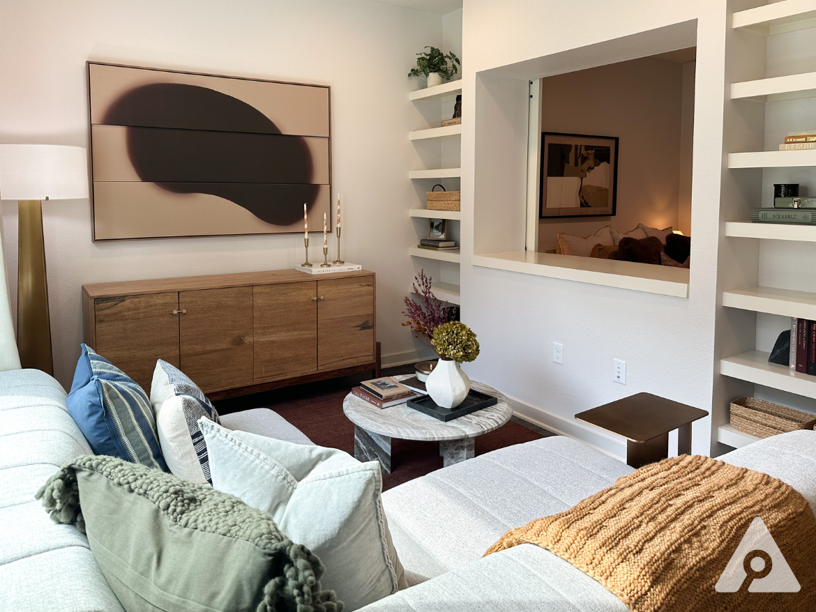 A cozy living room features a light gray sectional sofa adorned with various textured pillows, including a green pom-pom throw. A round marble coffee table sits in front, topped with a decorative vase and books. Behind the sofa, a wooden sideboard holds candles and a piece of abstract art hangs above it. A small bronze side table is positioned nearby. The room has built-in shelves displaying books and decorative items, and a cut-out window leads to another space, adding depth to the layout. The overall color palette is warm and inviting.