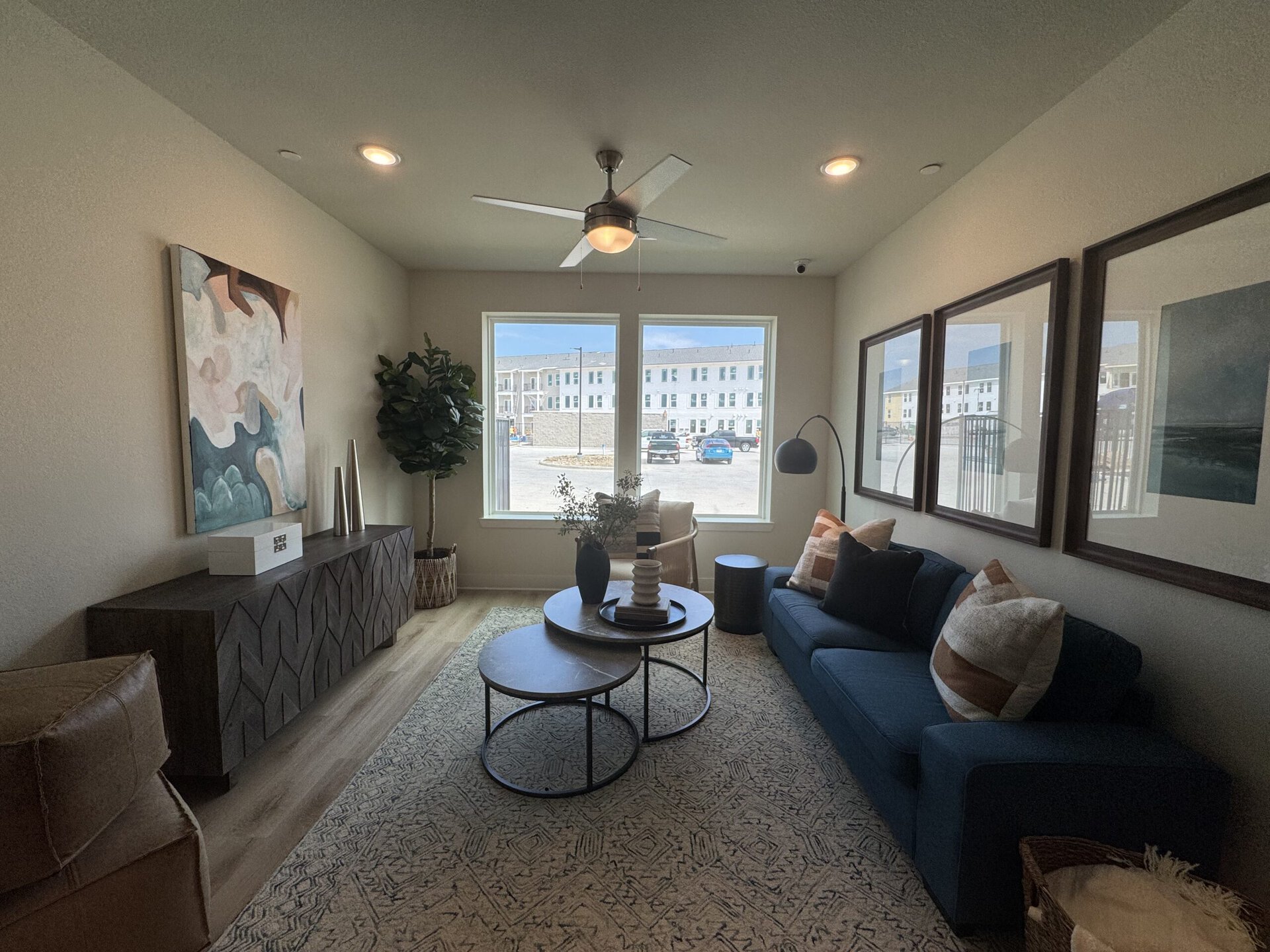 A modern living room features a blue sofa adorned with patterned throw pillows. In front of the sofa, there are two round coffee tables, one slightly nested within the other, topped with decorative items including a vase and a candle holder. A textured area rug covers the floor, and a dark wooden console table with a geometric design is positioned against the wall. Above the console, an abstract painting adds a splash of color. A tall potted plant stands beside a large window that lets in natural light, revealing a view of the outside area with parked cars. A ceiling fan and wall-mounted lamp complete the cozy atmosphere.