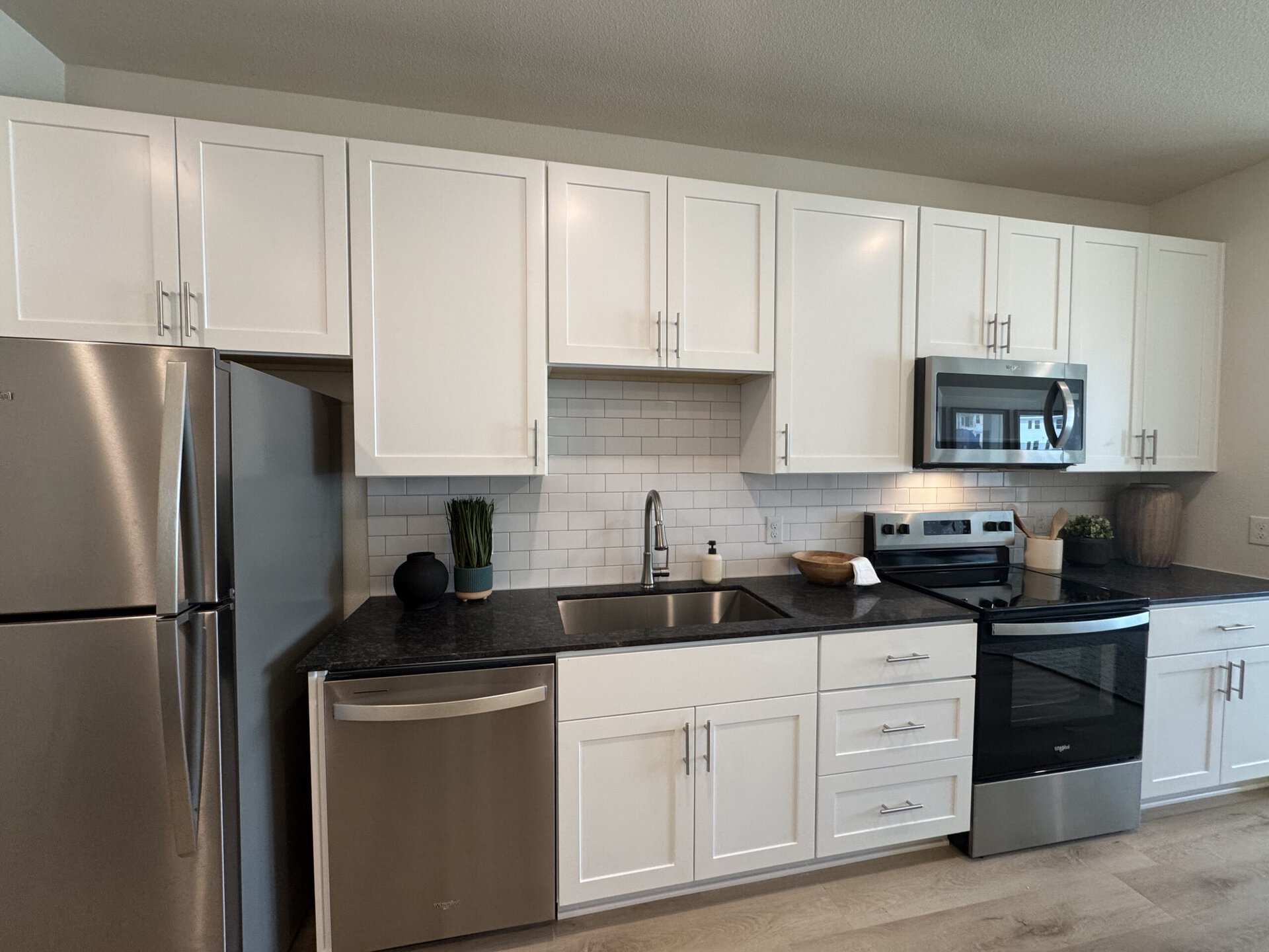 A modern kitchen features white cabinetry with sleek handles, a stainless steel refrigerator, and a dishwasher. The countertop is dark granite, complementing the white subway tile backsplash. A stainless steel sink with a pull-down faucet is positioned next to a black stove and microwave. Decorative elements include a small potted plant, a black vase, and a wooden bowl on the counter. The overall design is clean and contemporary.