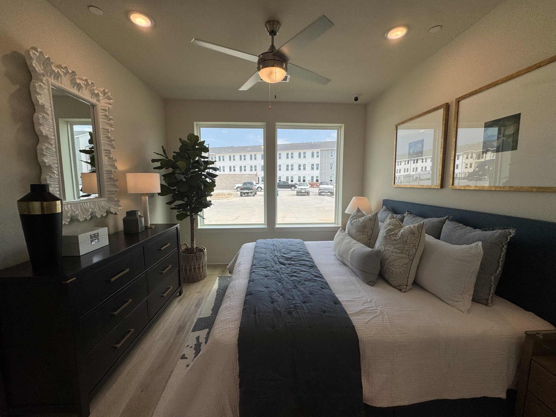 A cozy bedroom features a large bed with a dark blue headboard and multiple decorative pillows. A light gray blanket runs across the bed. To the left, a dark dresser holds a decorative mirror with a textured white frame, a black vase, a gray vase, and a small white box. A floor lamp with a white shade stands beside a faux plant in a woven basket. Two large windows allow natural light to fill the room, showcasing a view of a parking area and nearby buildings. A ceiling fan is visible above.