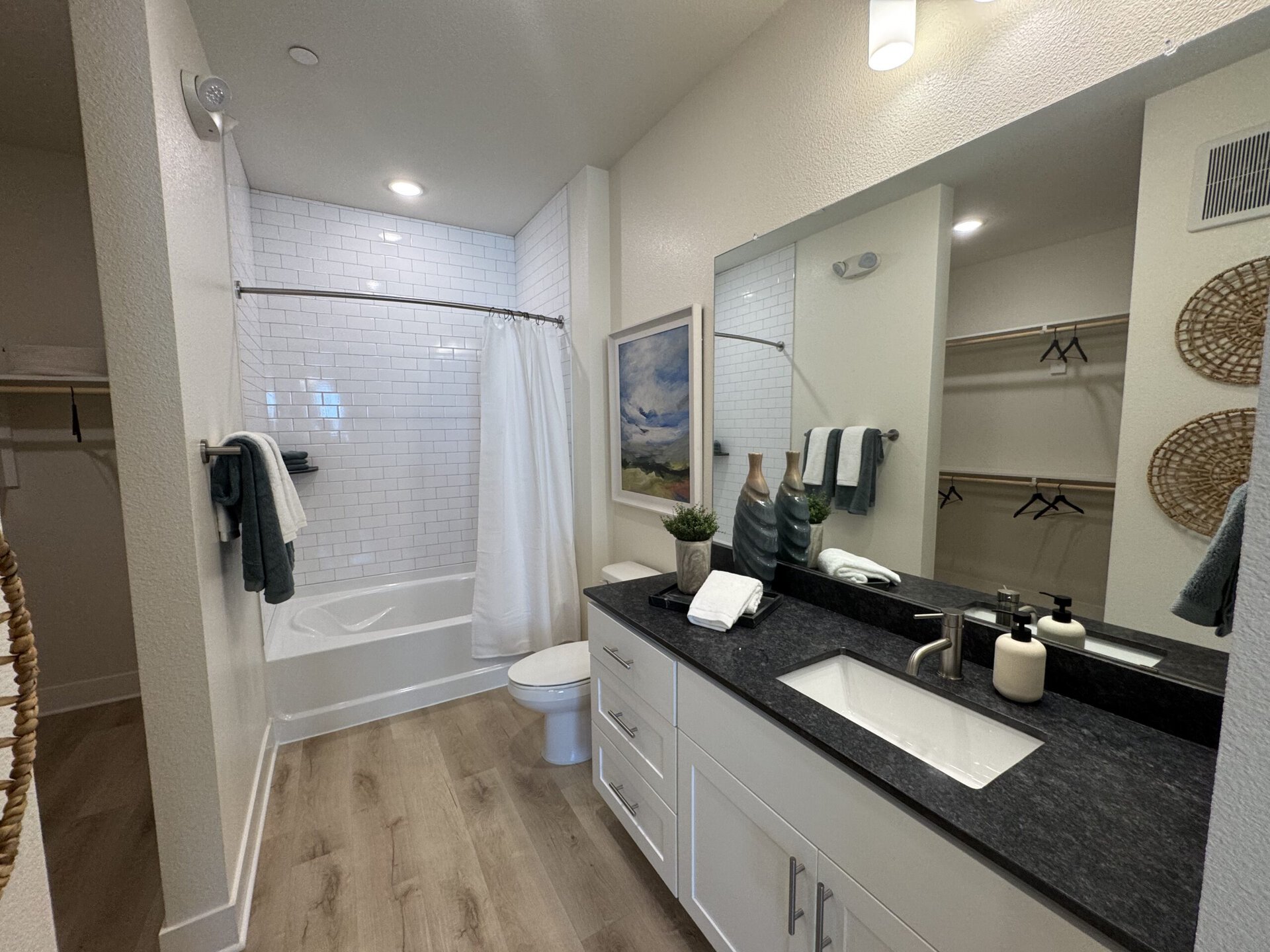 A modern bathroom features a white bathtub with a shower curtain, surrounded by white subway tiles. A sleek black countertop holds a sink, two decorative bottles, and neatly arranged towels. Above the sink, a large mirror reflects the space. To the side, a small plant adds a touch of greenery. A closet with hanging space is visible, and the floor is finished with light wood planks. The overall color scheme is neutral with accents of gray and white.