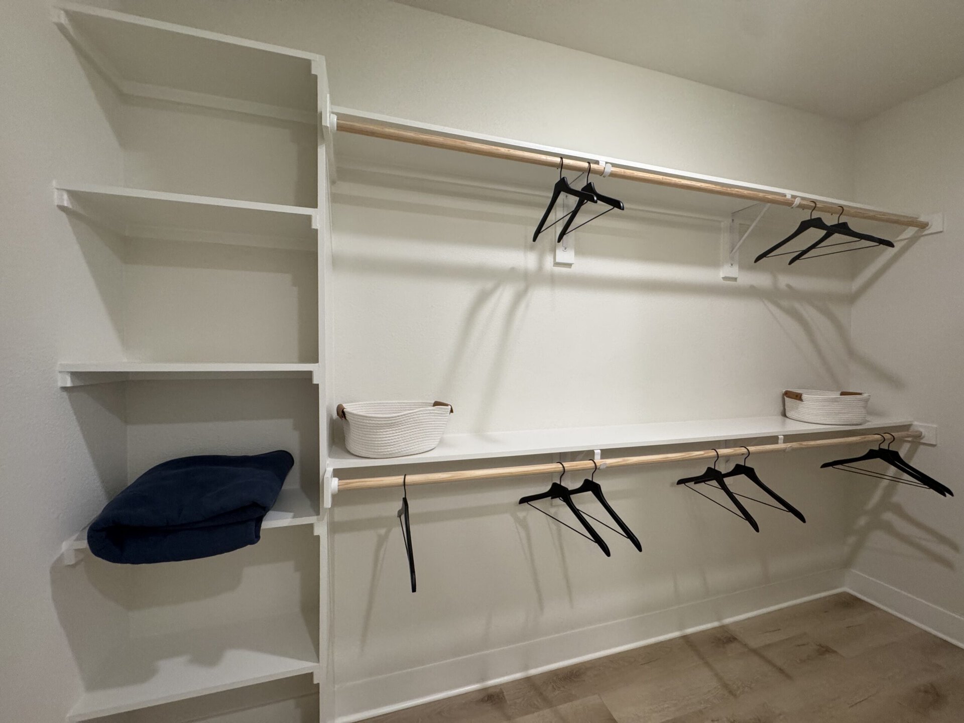 A minimalist closet space features a white wall-mounted shelving unit with a wooden hanging rod. Several black hangers are suspended from the rod. To the left, there are two open shelves, one holding a folded navy blue item and the other empty. A white woven basket with brown handles sits on the shelf below the hanging rod. The overall design is clean and organized, emphasizing functionality.