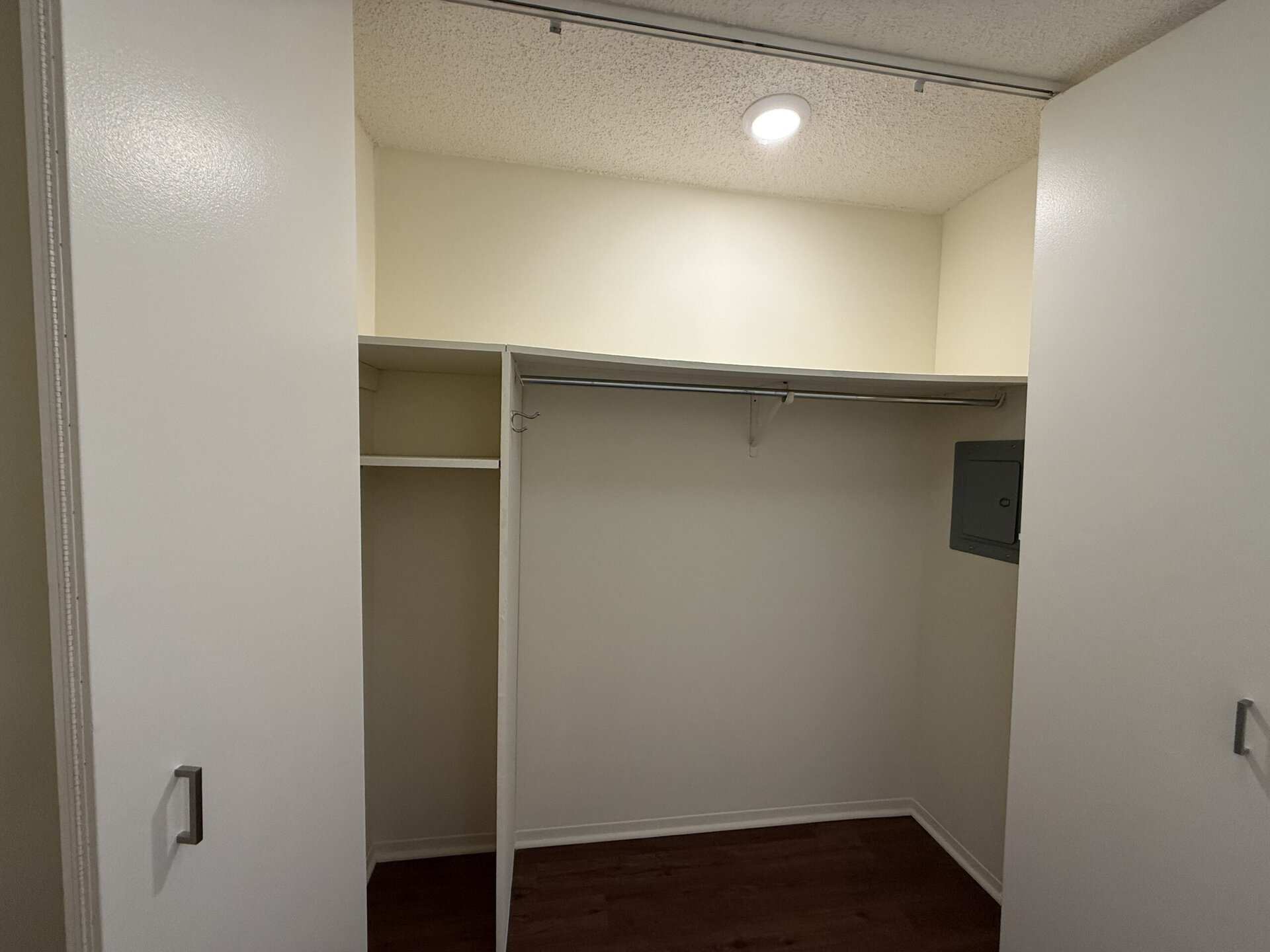 A spacious closet with white walls and a light fixture overhead. The closet features a hanging rod along the back wall and a shelf above it. To the left, there is an additional section with a shelf and no hanging rod. A small gray electrical panel is mounted on the right wall. The floor appears to be a wooden laminate.