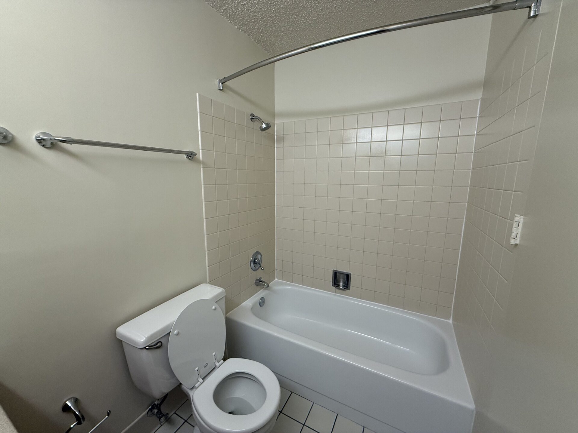 A clean, simple bathroom featuring a white bathtub with a chrome faucet and a showerhead. The walls are tiled in light beige squares, and there are two chrome grab bars mounted on the wall. A white toilet is positioned next to the bathtub. The overall color scheme is neutral, with a light-colored wall adjacent to the tiled area.