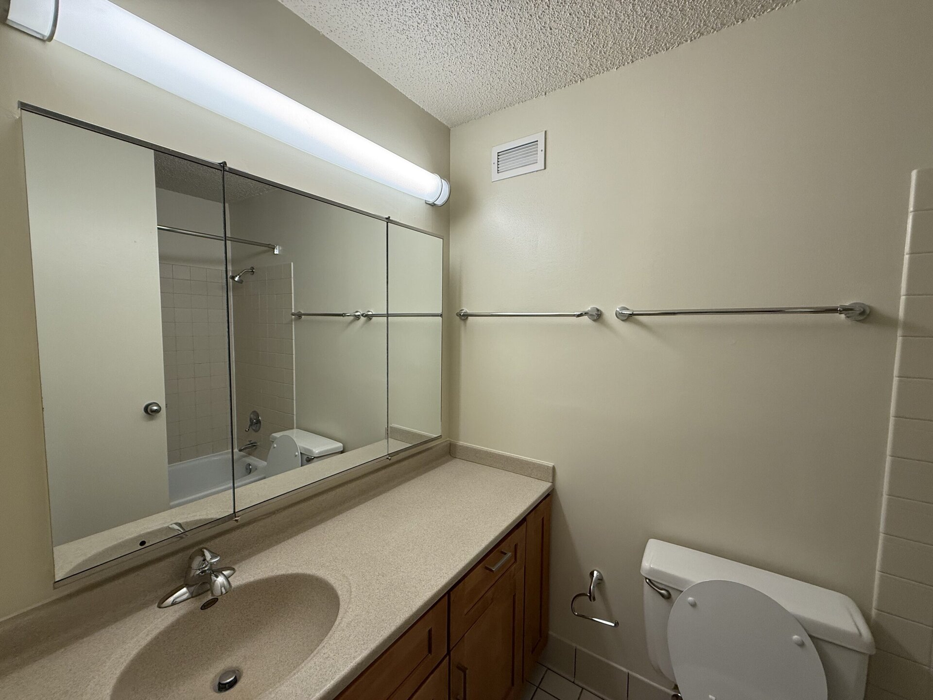 A bathroom with a light-colored wall and a large mirror spanning the countertop. The countertop is beige and features a sink with a faucet. To the right, a white toilet is visible, and in the background, a bathtub with a showerhead is partially seen. Two towel bars are mounted on the wall above the countertop, and a ventilation grille is located near the ceiling. The overall space appears clean and well-lit.