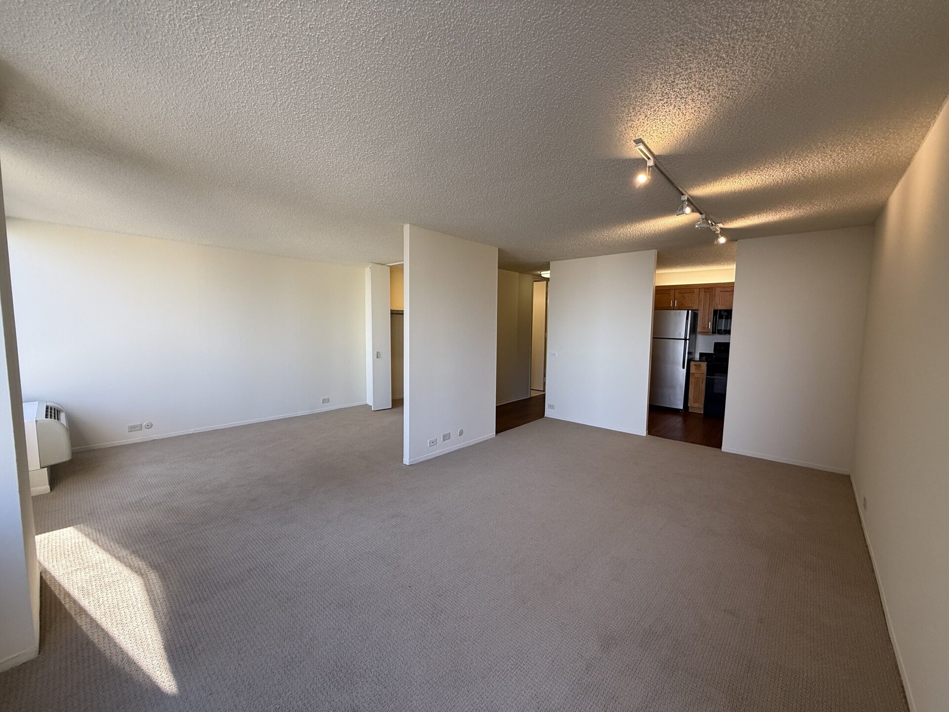 A spacious, empty living area with beige carpet and white walls. On the left, there is a small air conditioning unit. To the right, a doorway leads to a kitchen area featuring wooden cabinets and stainless steel appliances, including a refrigerator. The room is well-lit with natural light coming from a window, and track lighting is installed on the ceiling. The layout suggests an open floor plan, with a clear division between the living space and the kitchen.