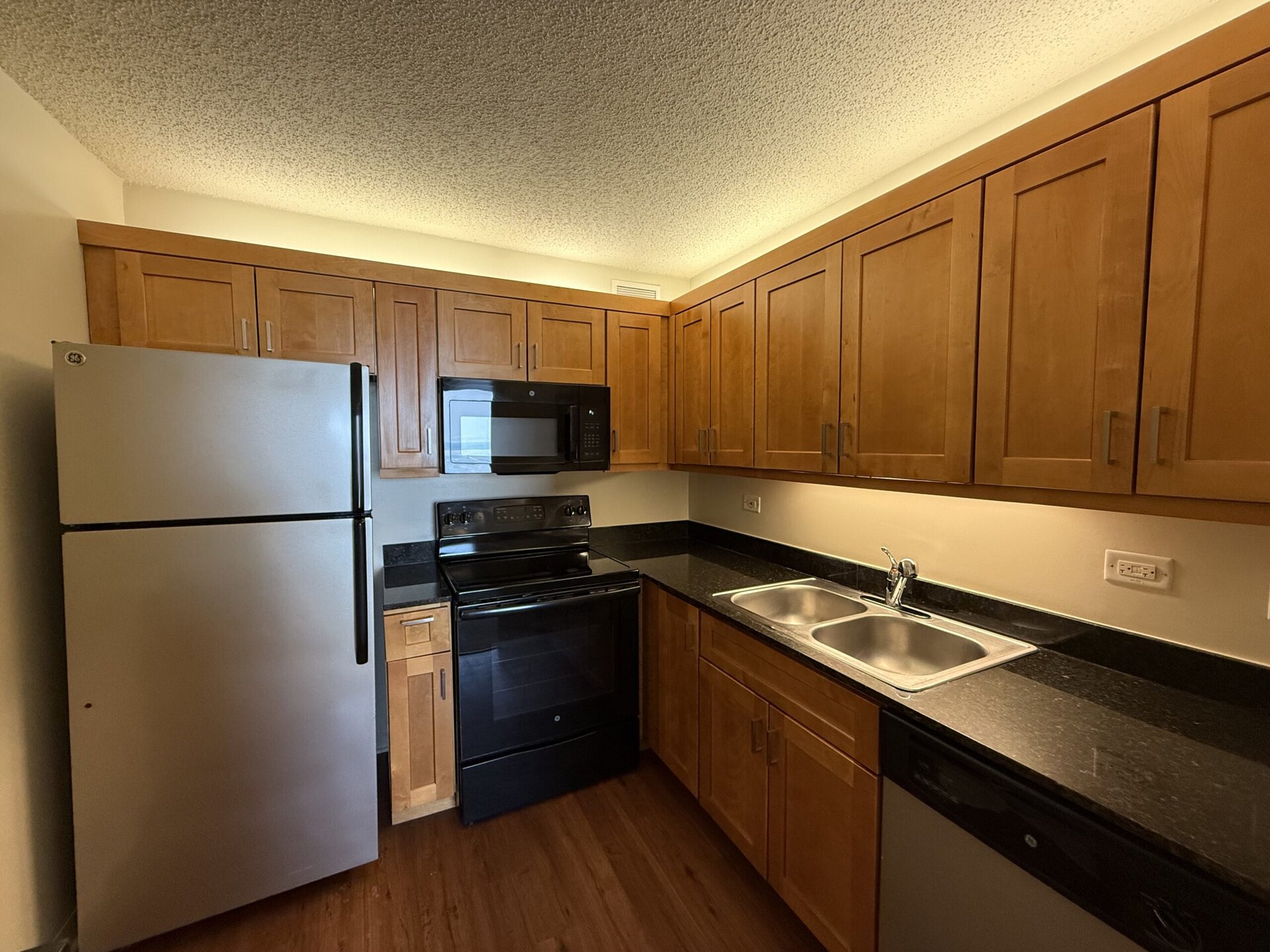 A modern kitchen featuring wooden cabinets with a warm finish, a black countertop, and stainless steel appliances. The appliances include a silver refrigerator, a black oven, and a microwave mounted above the stove. There are two stainless steel sinks with a faucet, and the kitchen has a well-lit atmosphere with overhead lighting. The flooring appears to be a dark wood.